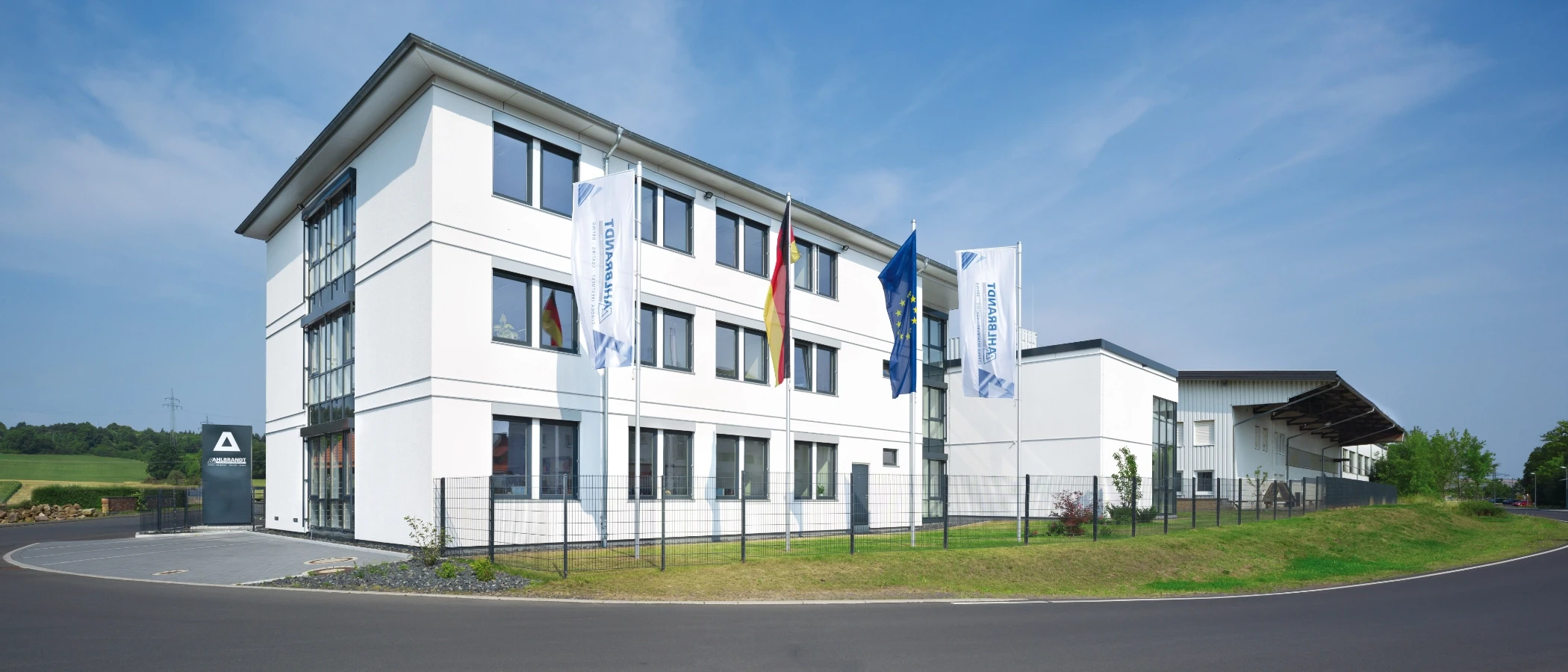 Modernes weißes Bürogebäude mit Fensterfronten, deutscher und EU-Flagge sowie Werbeschild vor blauem Himmel.