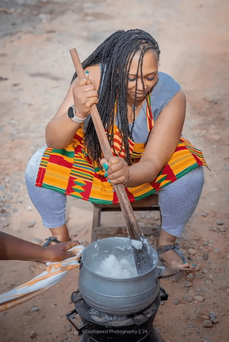 A traveler uses a traditional wooden stick to cook banku in a metal pot.