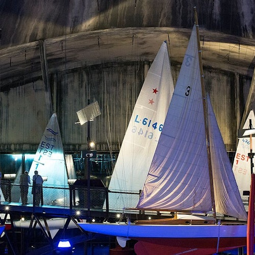 Beltéri vitorlásbemutató megvilágított vitorlákkal, amelyek különböző mintákat és számokat ábrázolnak. A háttérben sziluettként feltűnő alakok láthatók.