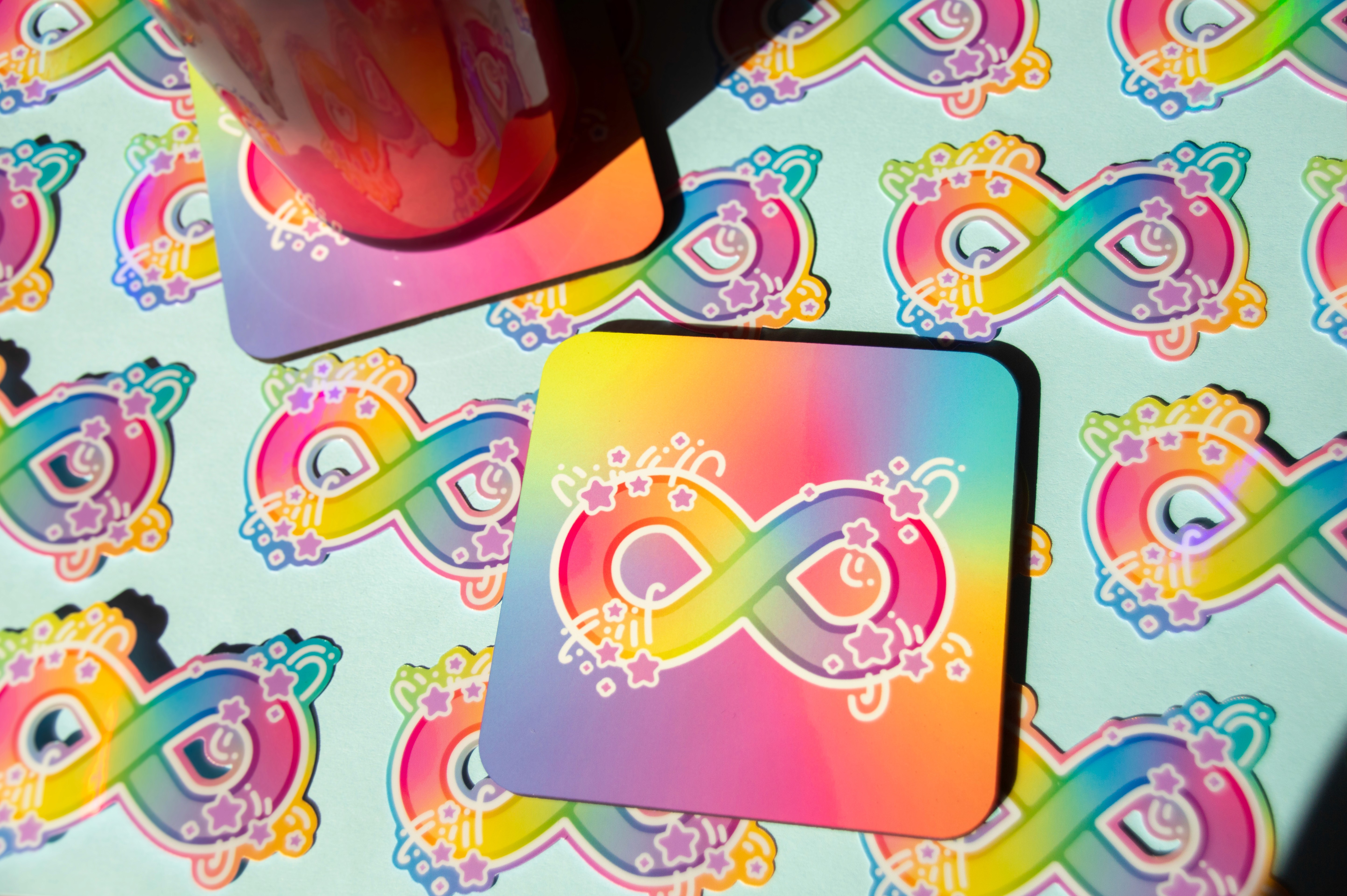 A rainbow coloured drinks coaster with an infinity symbol with stars surrounding it. The infinity symbol design is also on stickers that surround the coaster.