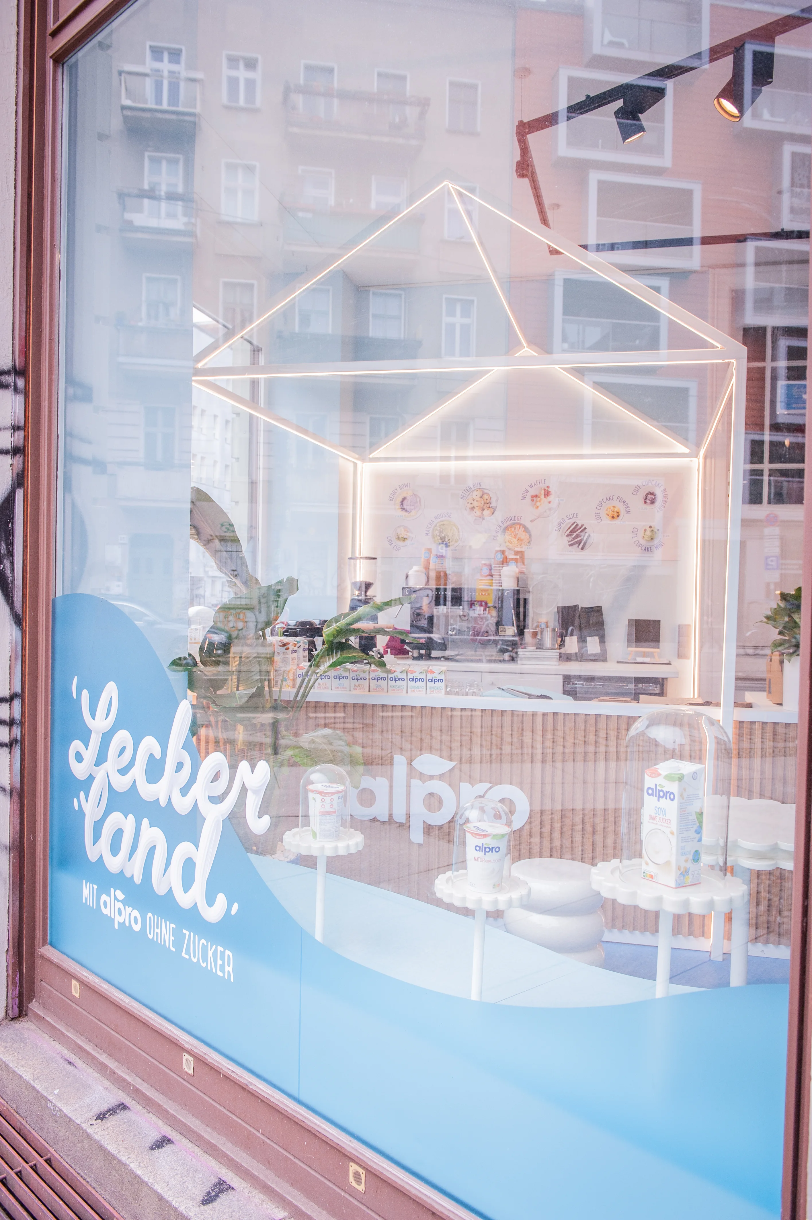 Shop window of the Alpro Leckerland pop-up with blue wave decal and view of the illuminated house-shaped counter.