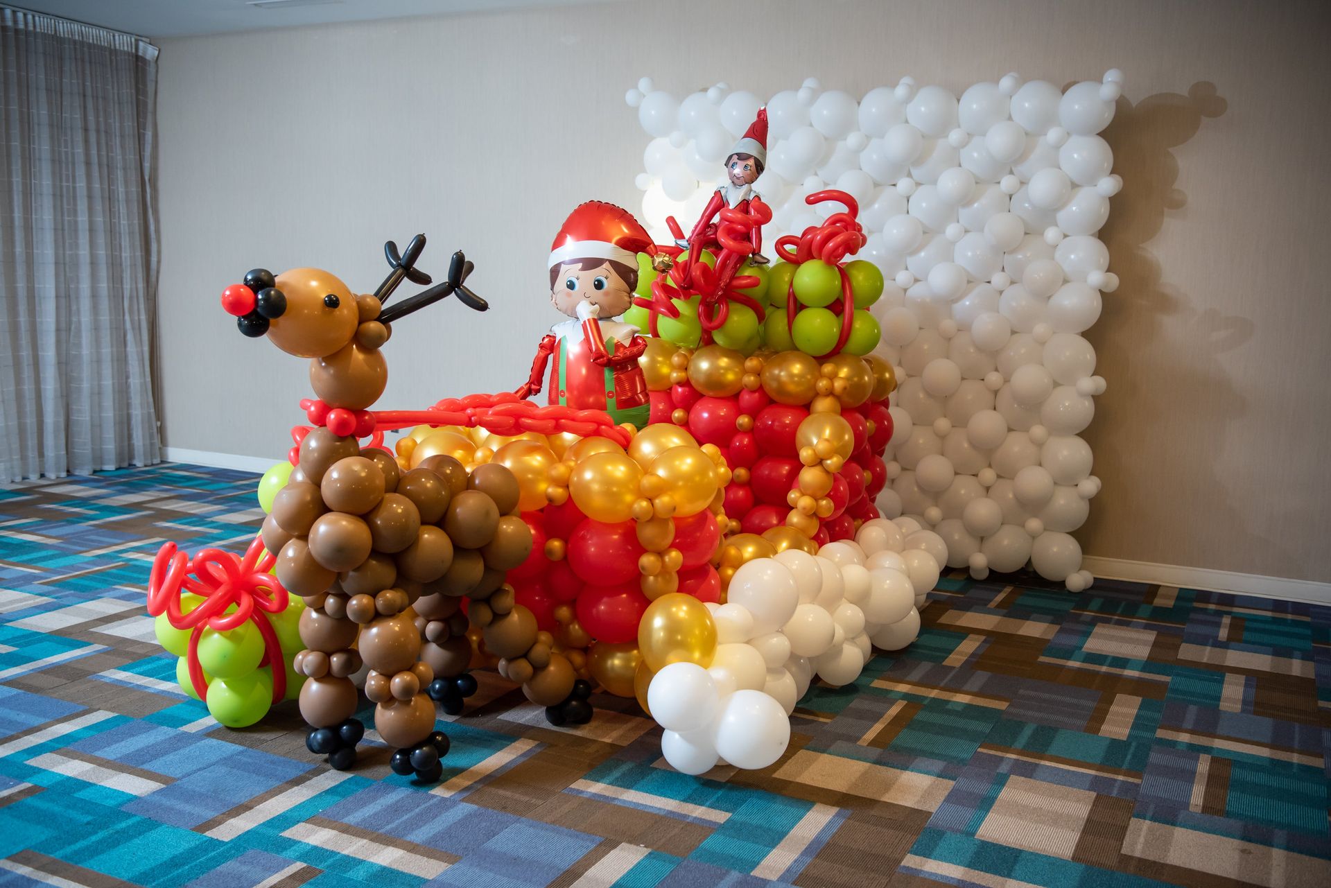 A colorful balloon sculpture depicts a festive Christmas scene with a reindeer pulling a sleigh filled with gifts and two cheerful elves, all crafted from variously colored balloons, sitting in a bright, contemporary room.