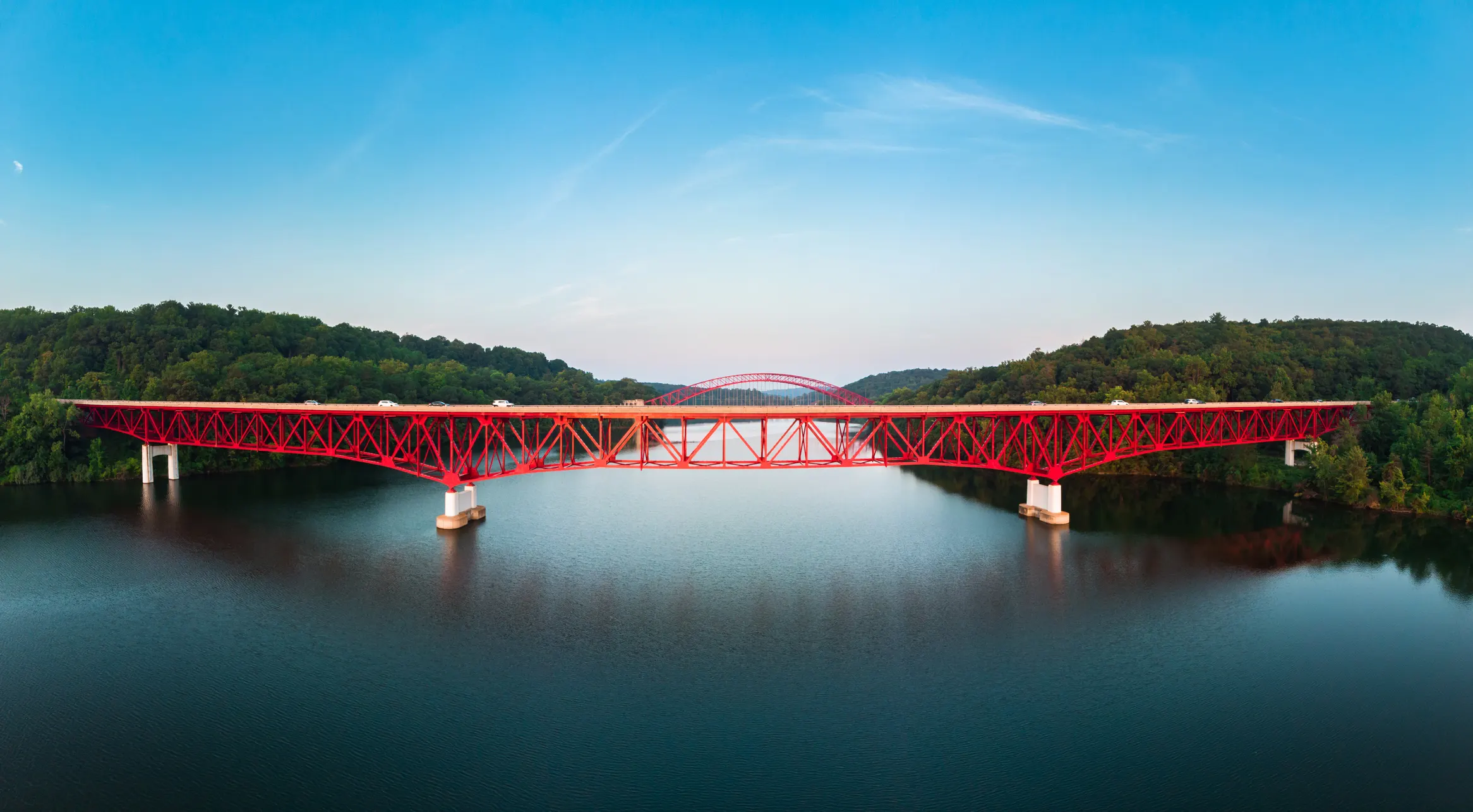 A red steel bridge spanning a calm river surrounded by green hills at sunset — symbolizing GIA Group, LLC’s role in helping new trucking businesses build strong foundations and move forward with confidence.