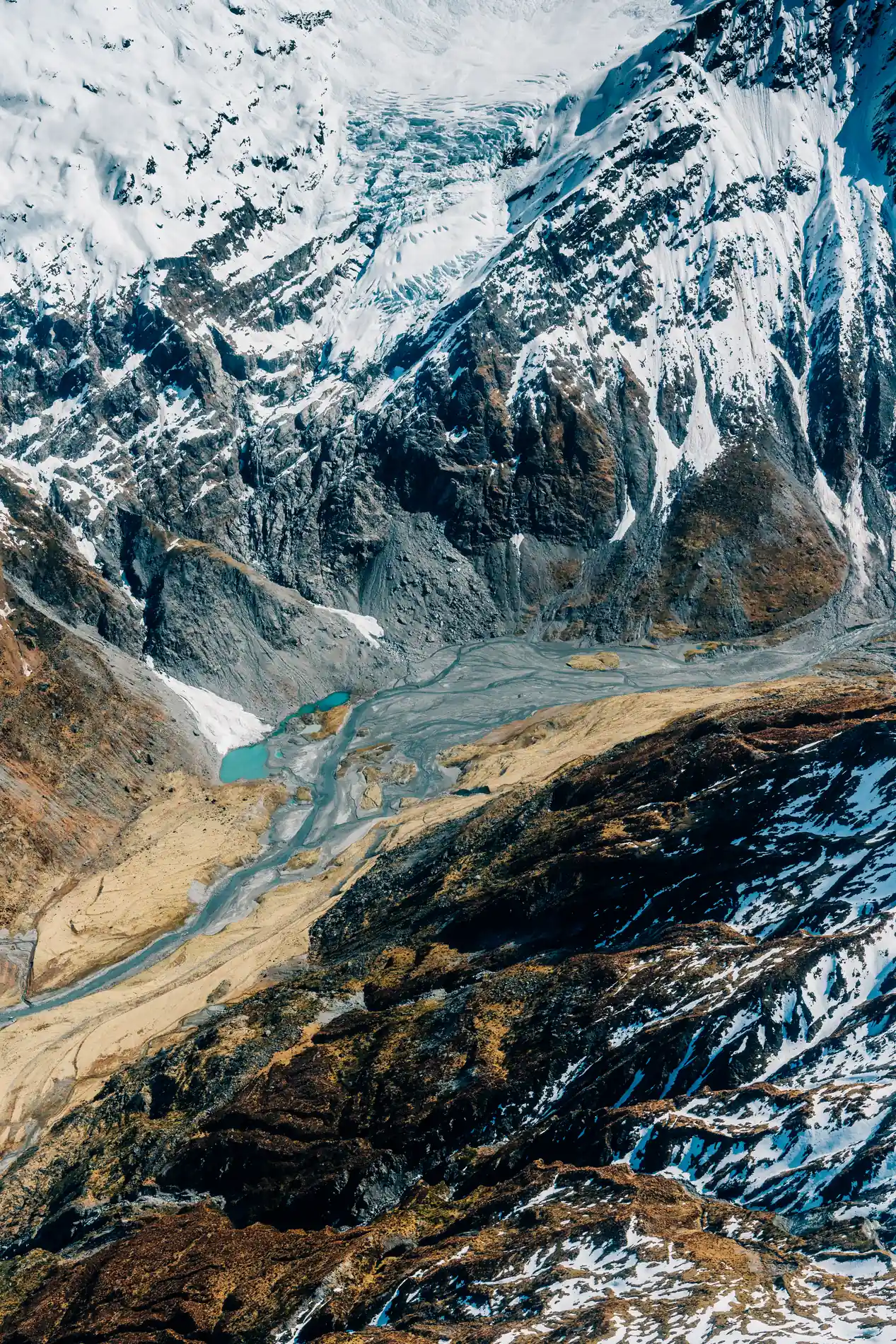 Aerial of a glacial lake and meltwater channels below steep snow-streaked slopes