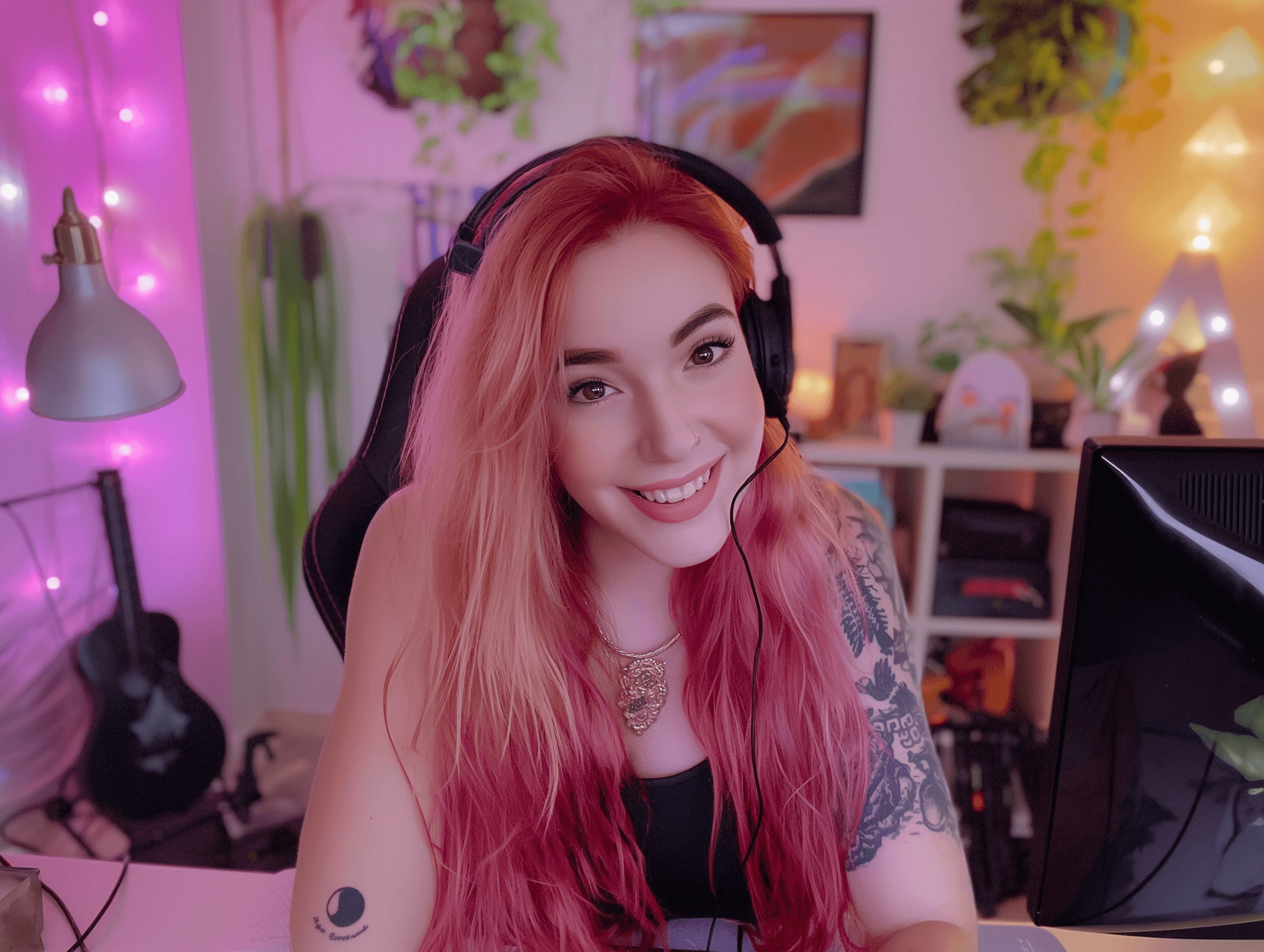 A smiling person with long pink hair sits at a desk decorated with plants and colorful lights, wearing headphones.