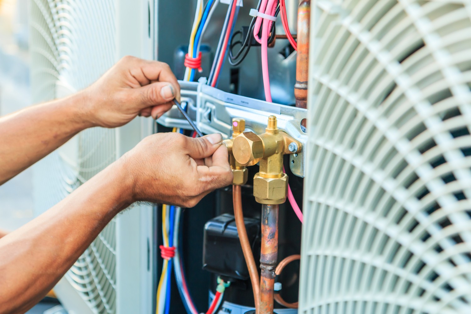 A person is adjusting electrical wires and components inside an HVAC unit. Their hands are visible working with tools to connect or manipulate the colorful wires and mechanical parts inside the unit. This scene, typical of AC repair in Asheville, also shows the unit's fan and other components partially visible.