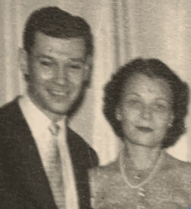 Black and white portrait of a smiling man and woman from the mid-20th century. The man wears a suit and tie with coiffed shorter hair and the woman has a relaxed updo pinned back and sports a chiffon and lace dress with a pearl necklace.