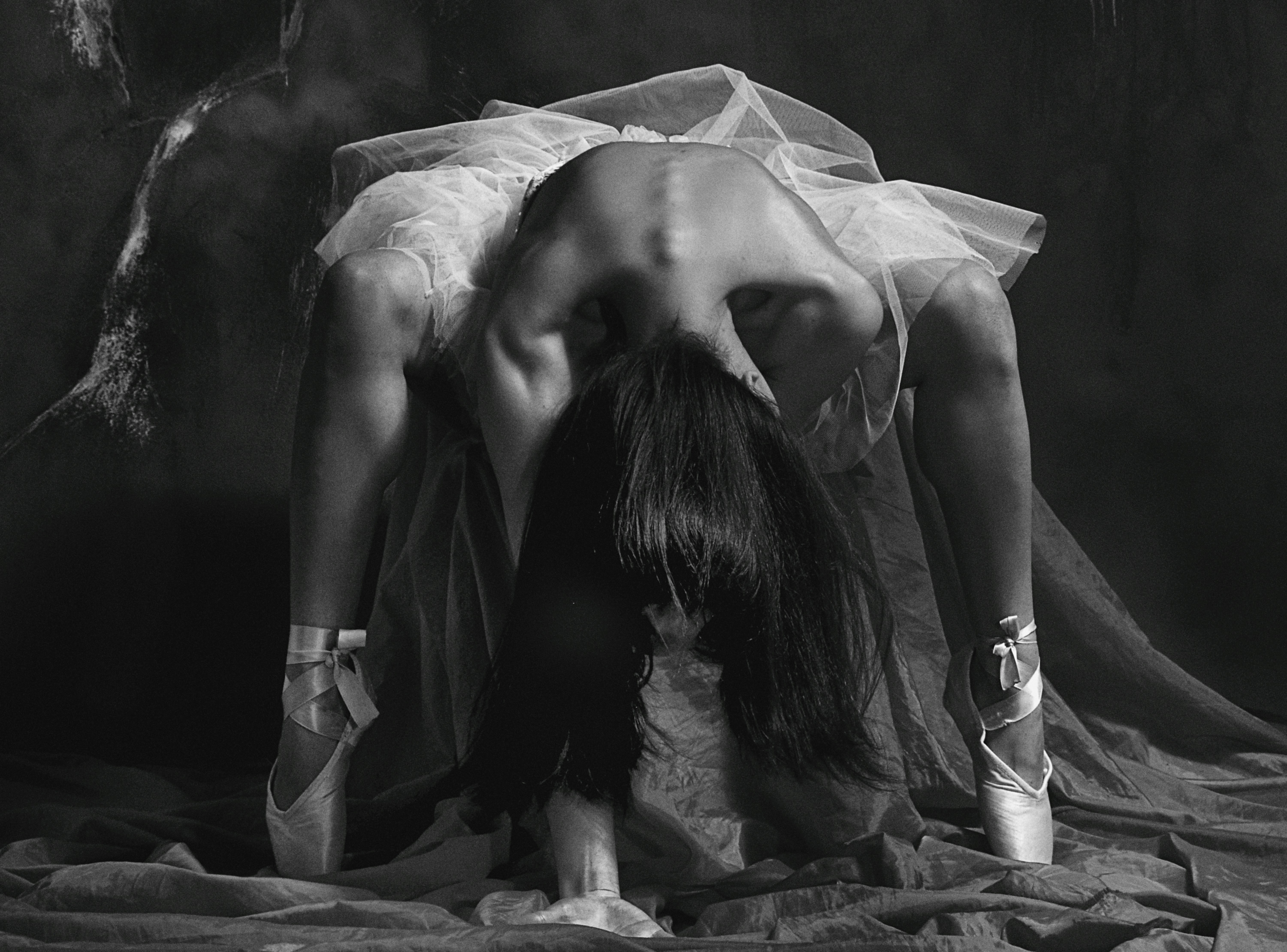 Black and white studio photography of a ballerina bent forward in pointe shoes, wearing a frayed white tulle tutu against a dark textured backdrop, with her face hidden.