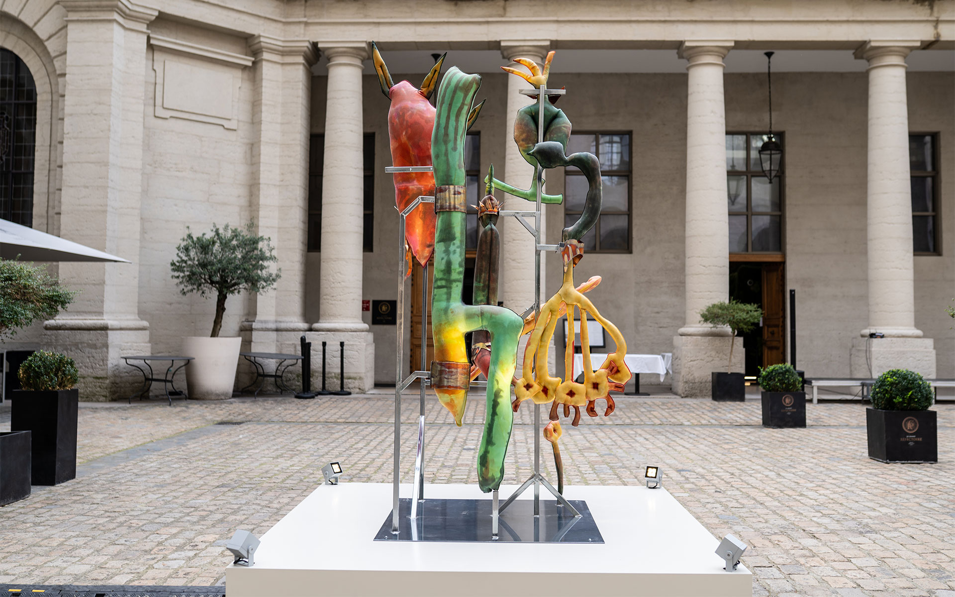Une sculpture abstraite et organique est exposée sur un socle blanc au centre d'une cour pavée entourée de colonnes classiques. L'œuvre est composée d'une structure métallique supportant plusieurs éléments aux formes courbes et aux textures variées, aux teintes vertes, jaunes, orangées et brunes, évoquant des formes végétales ou biologiques. Des petits projecteurs sont disposés autour de la base du socle, illuminant la sculpture dans cet espace architectural formel.