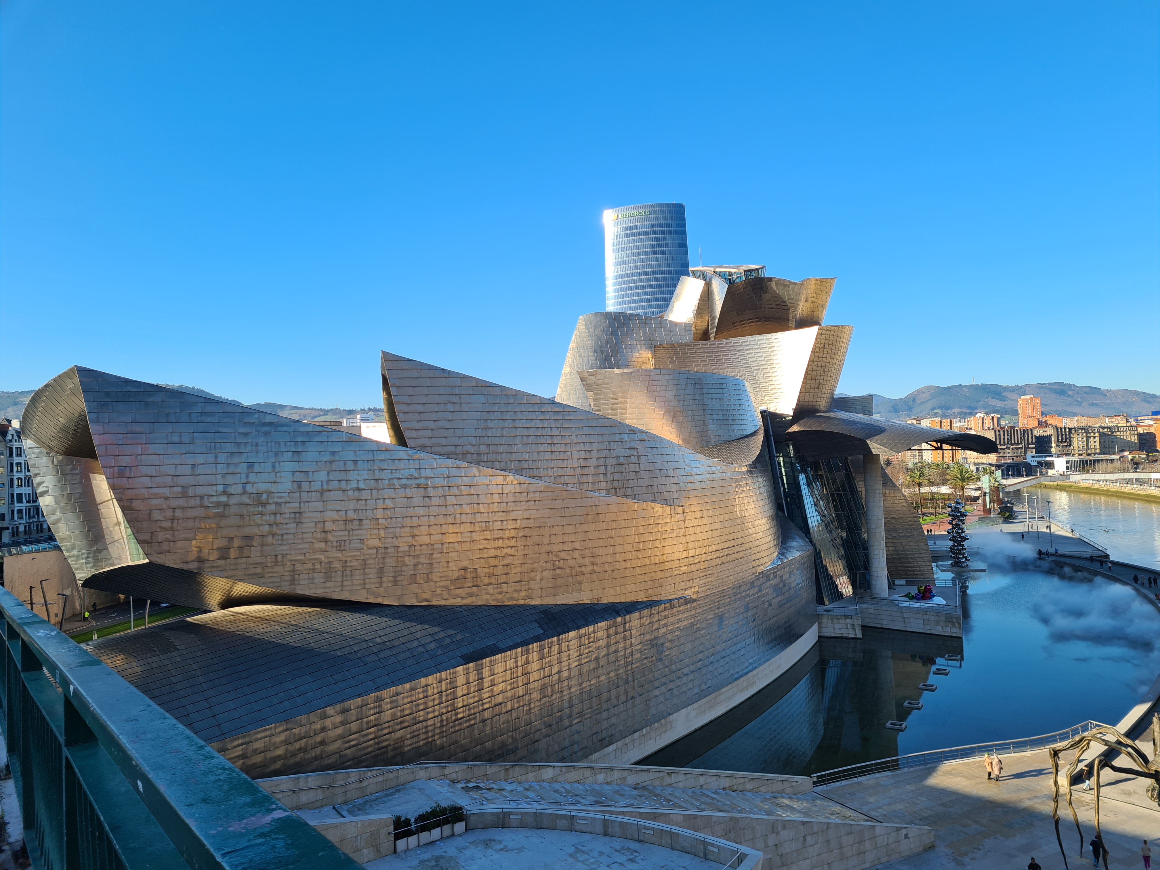 A person walks along a seaside pathway, gesturing with one hand, wearing a jacket and casual shoes.