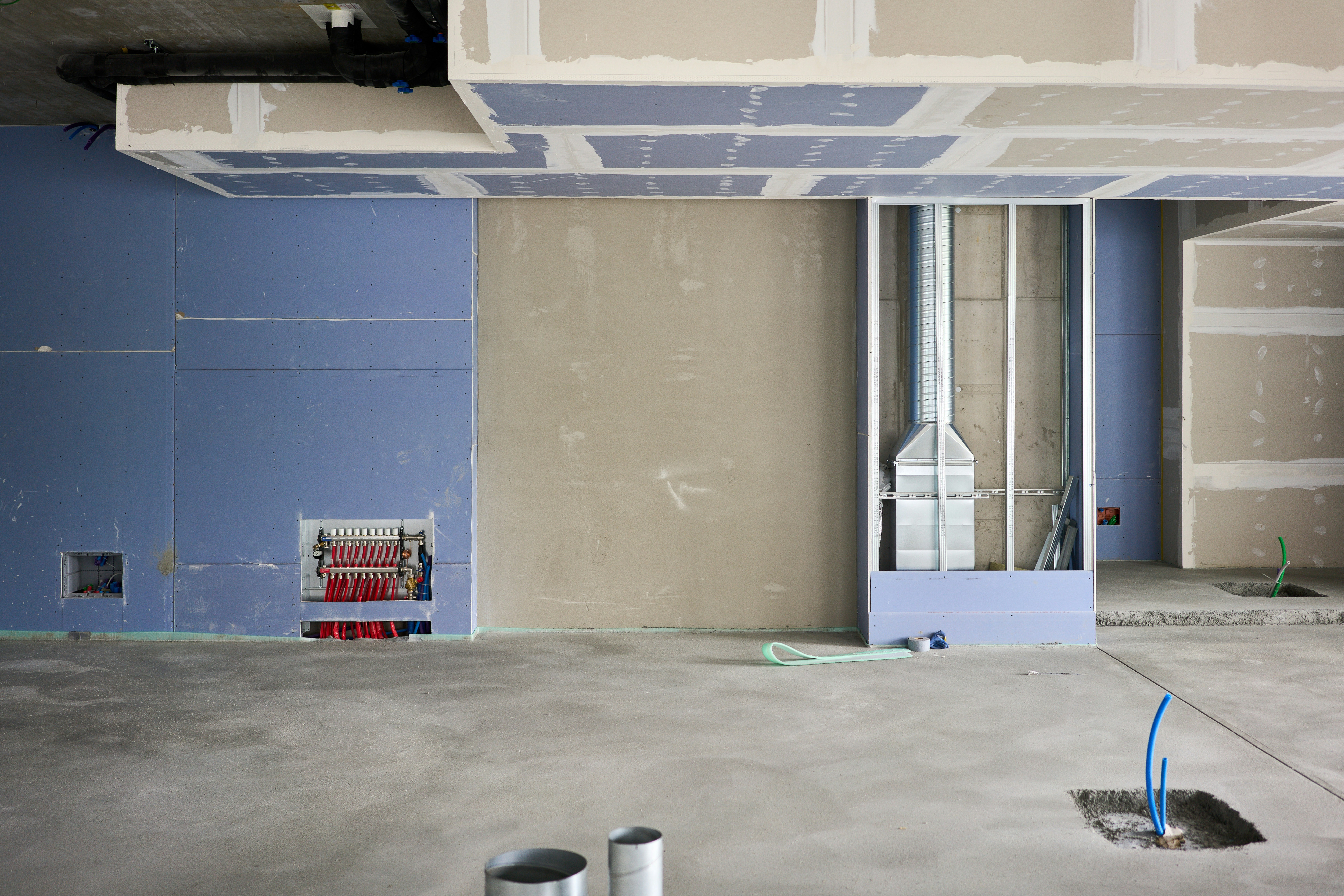 Intérieur d'un chantier de construction avec câbles apparents, conduits et cloisons sèches non finies. Des tuyaux et des câbles émergent du sol en béton, indiquant que les travaux sont en cours.