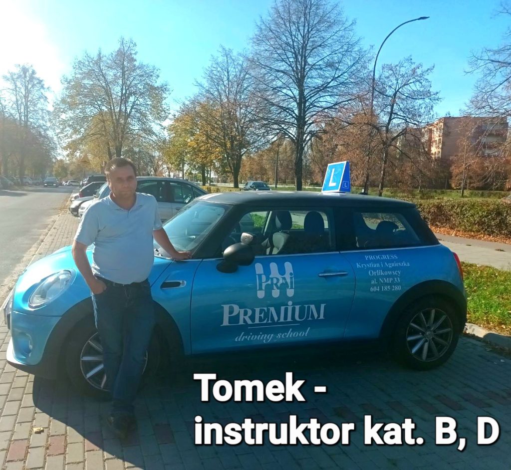 Instruktor Tomek pozuje w słońcu przy samochodzie na tle drzew i zabudowy.