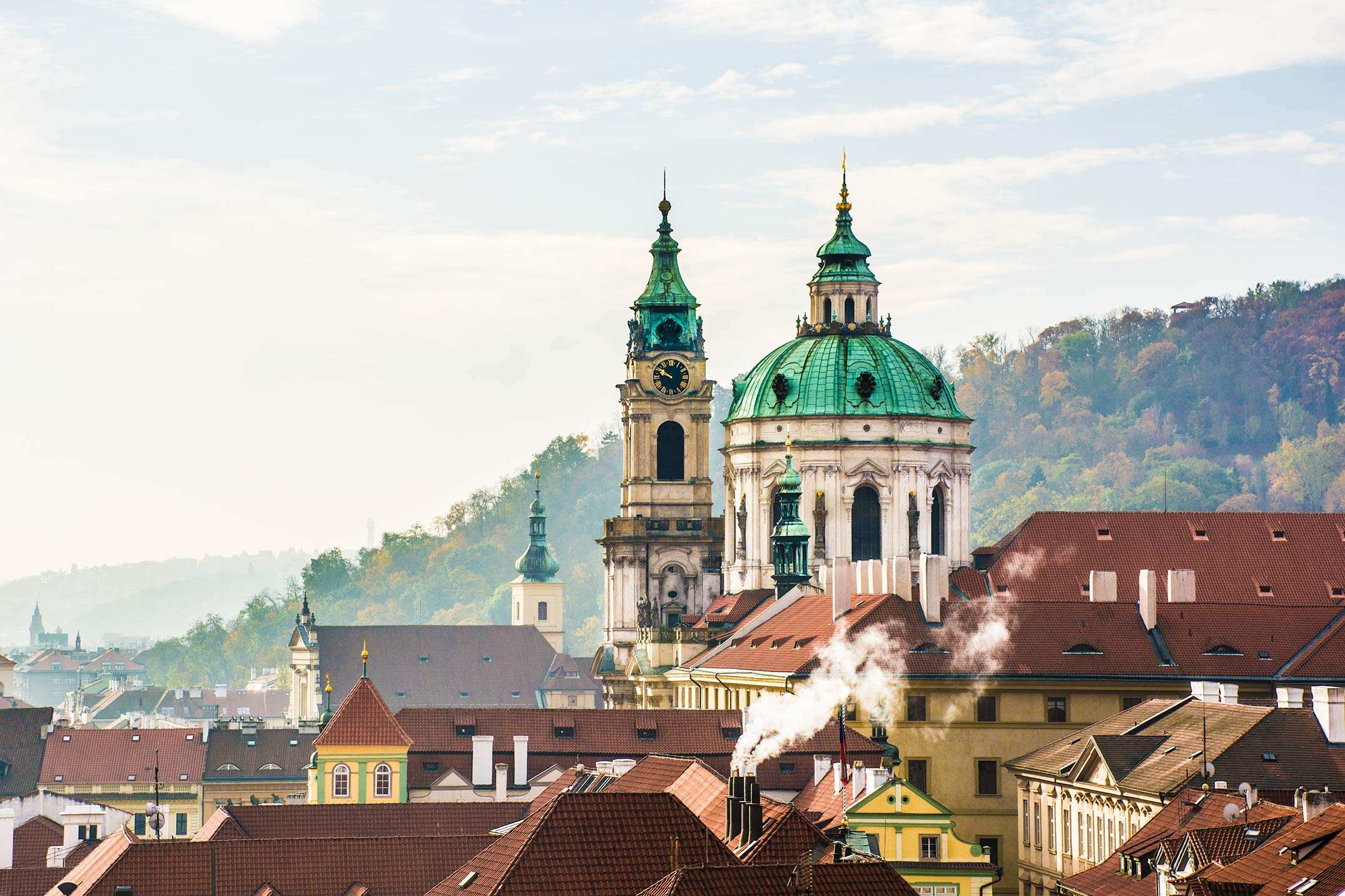A panoramic view of a European town with red-roofed buildings and two prominent green-domed churches under a blue sky.