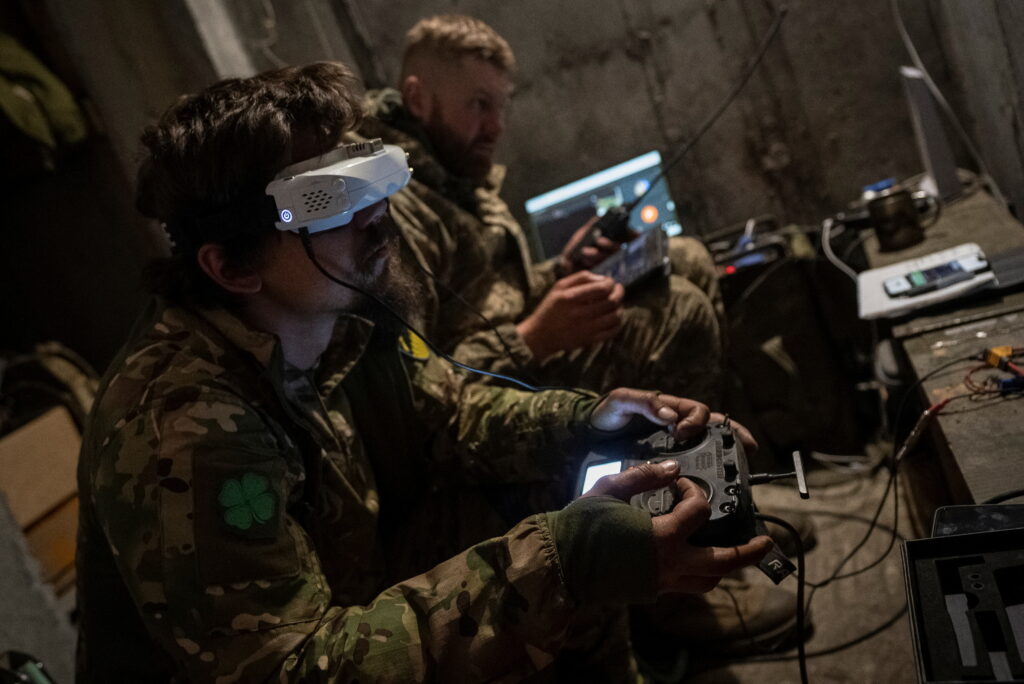 Two soldiers seated indoors, operating a drone using VR headsets and controllers during a military exercise. The image depicts the use of virtual reality (VR) technology for drone piloting and tactical simulations, emphasizing its role in VR training and battlefield operations. This scene illustrates the integration of VR development into military settings for enhanced precision and operational efficiency.