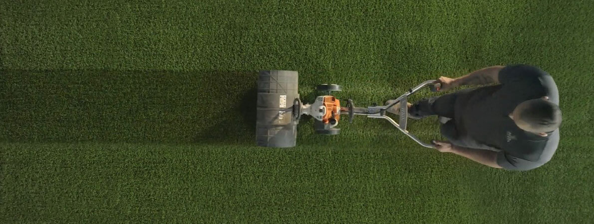 Pictured from above, a man uses a power broom to maintain the appearance of synthetic grass.