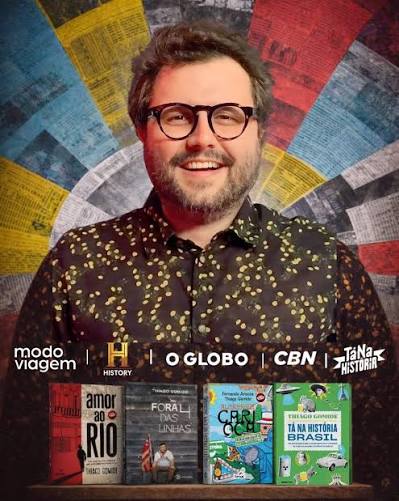 Man with glasses and patterned shirt smiling. Co-criador do Tá na História. Thiago Gomide. Fernanda Arouca