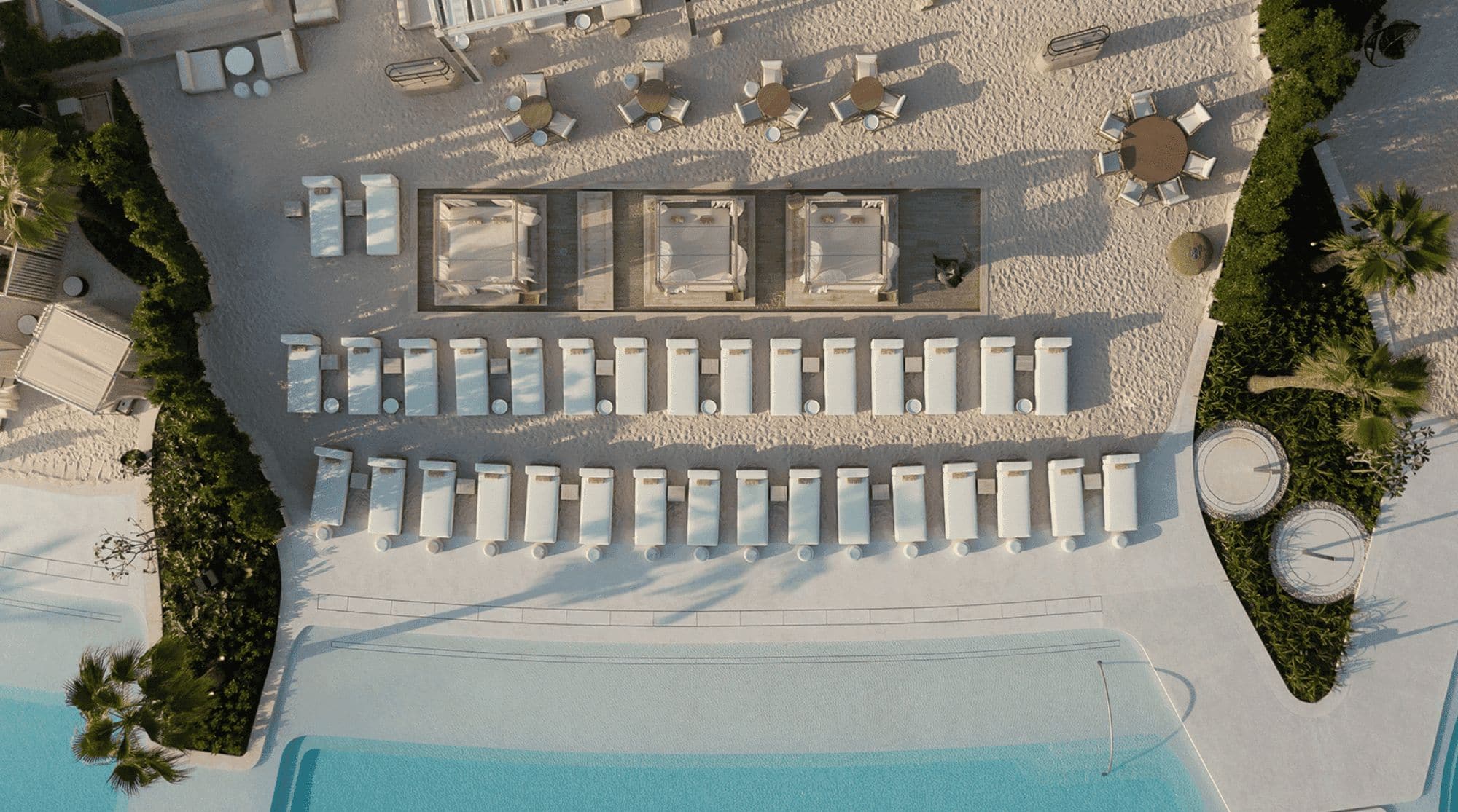 An aerial view of a beachfront pool area with neatly arranged sun loungers, cabanas, and sandy surroundings.