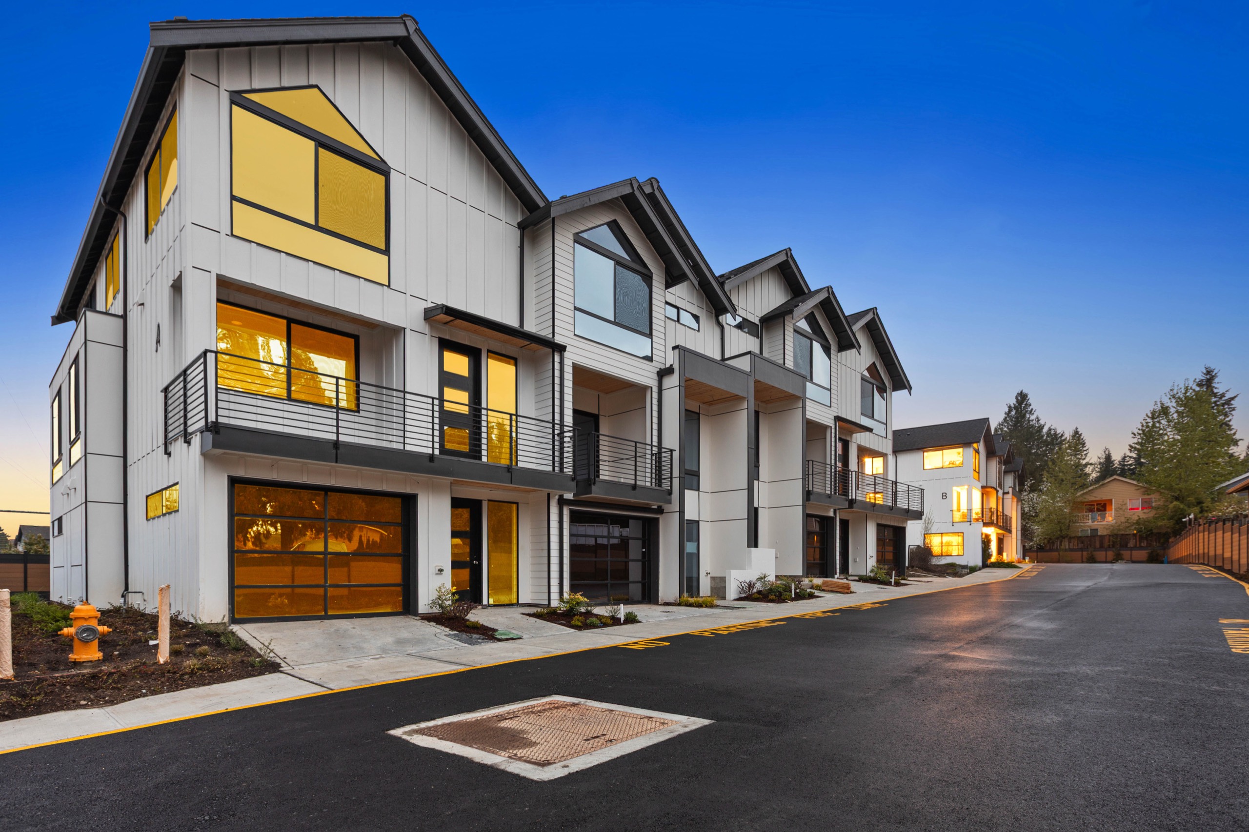 Twilight exterior of a modern luxury townhome complex with white board-and-batten facade, two-car garages, upper-floor balconies, and warm interior lighting
