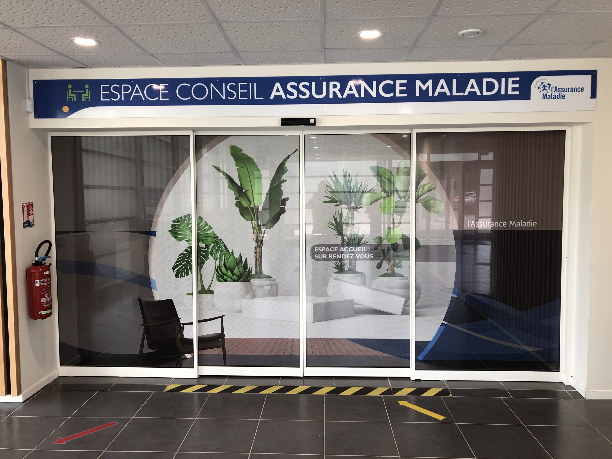 Portes coulissantes en verre surmontées d'une enseigne « Espace Conseil Assurance Maladie », agrémentées de plantes d'intérieur et de chaises, créant une atmosphère calme et accueillante.