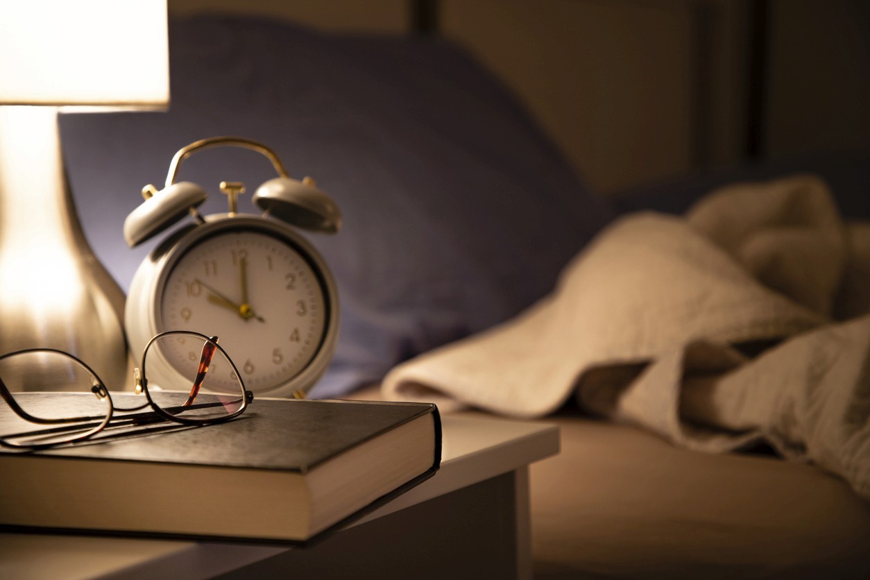 A bed side table with an alarm clock, book and reading glasses. To the back is a bed with disturbed sheets.
