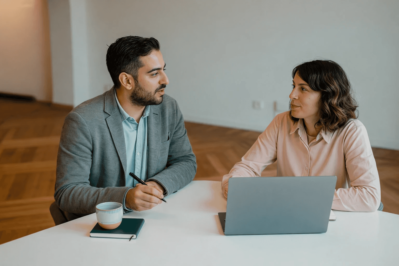 Two colleagues talking across a table with a laptop and coffee cup