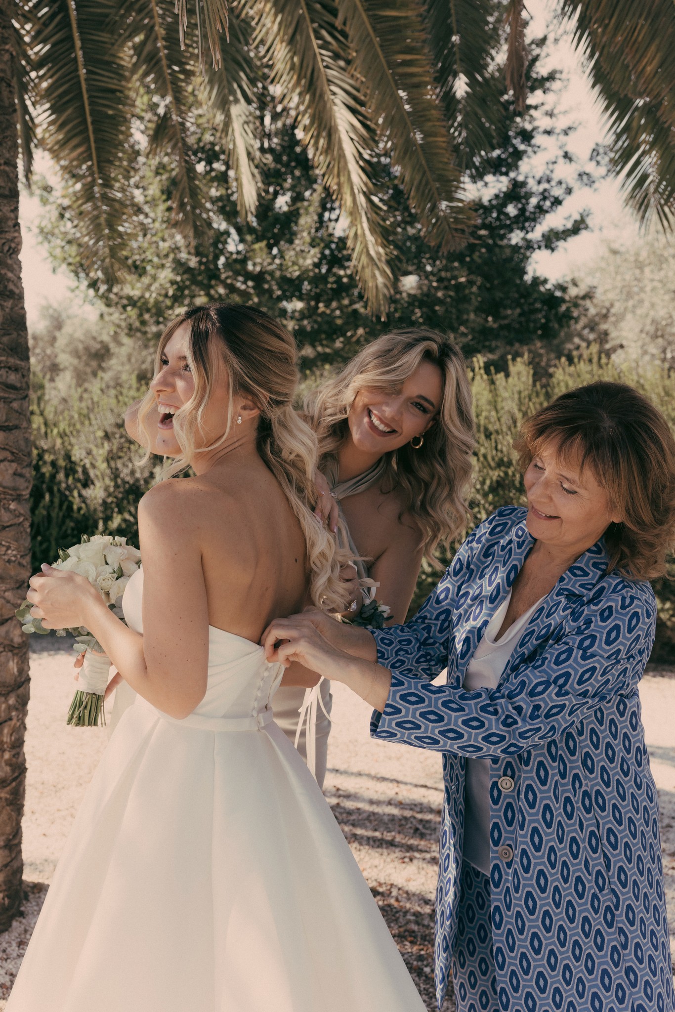 Sposa in abito bianco sorride tra la madre e un'amica, preparativi matrimoniali a Bolgheri tra i cipressi toscani