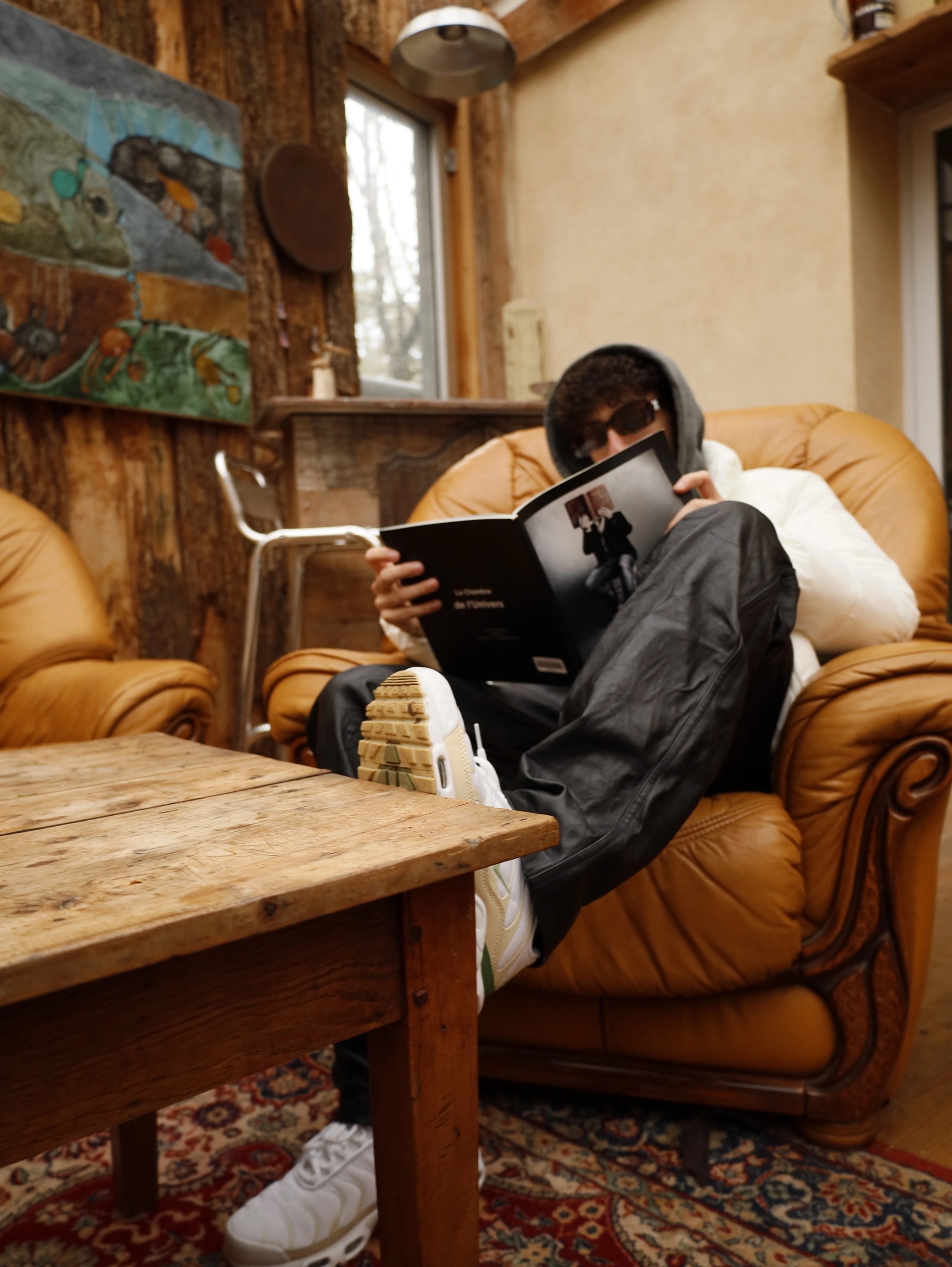 A person sits comfortably on a leather couch, sipping a drink, with a child nearby, in a cozy room.