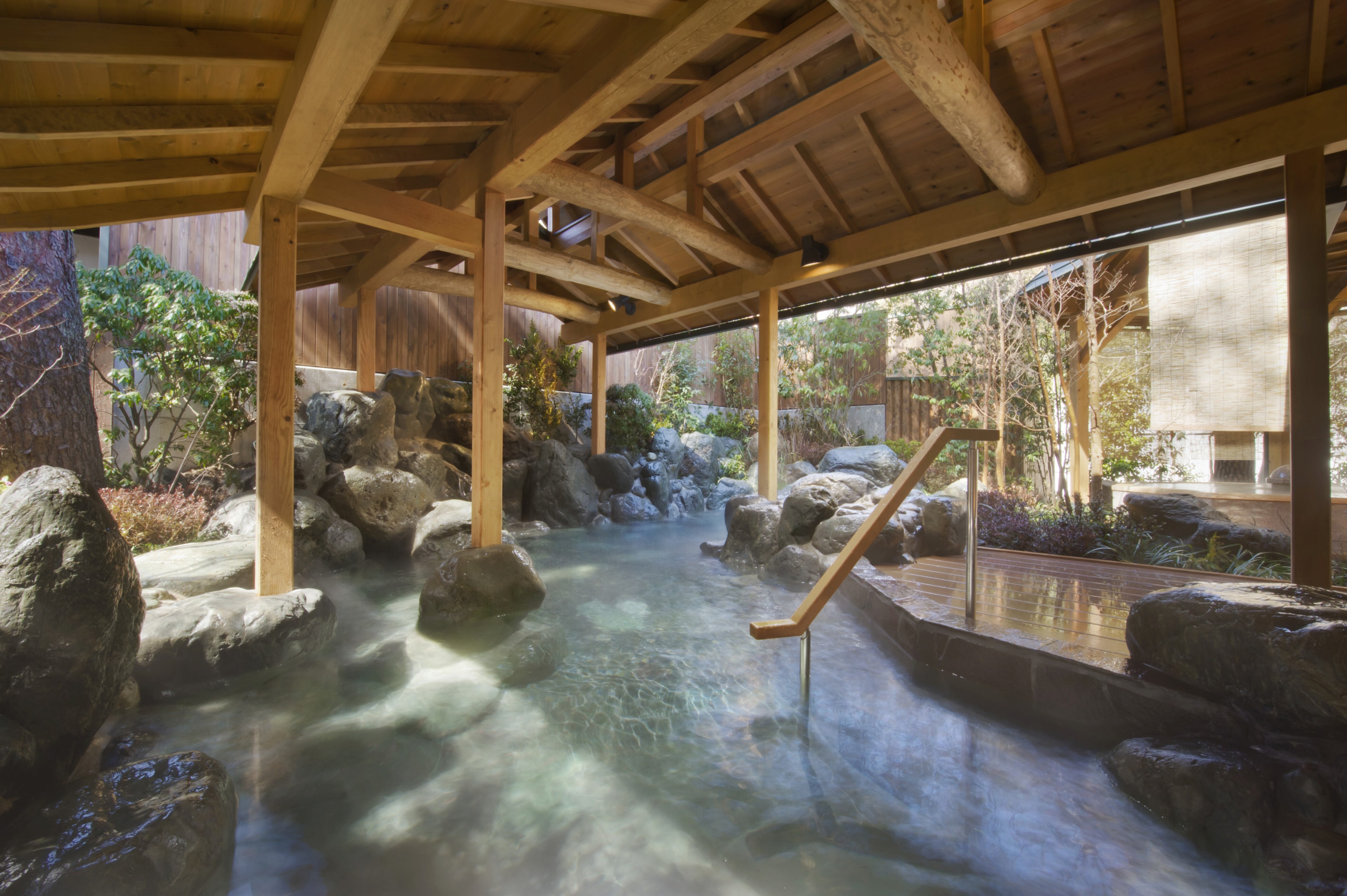 A serene outdoor Japanese hot spring (onsen) with a rock-lined pool of water, set under a traditional wooden pavilion with exposed beams. A small wooden spout pours water into the pool, which is surrounded by lush green plants and a stone wall.