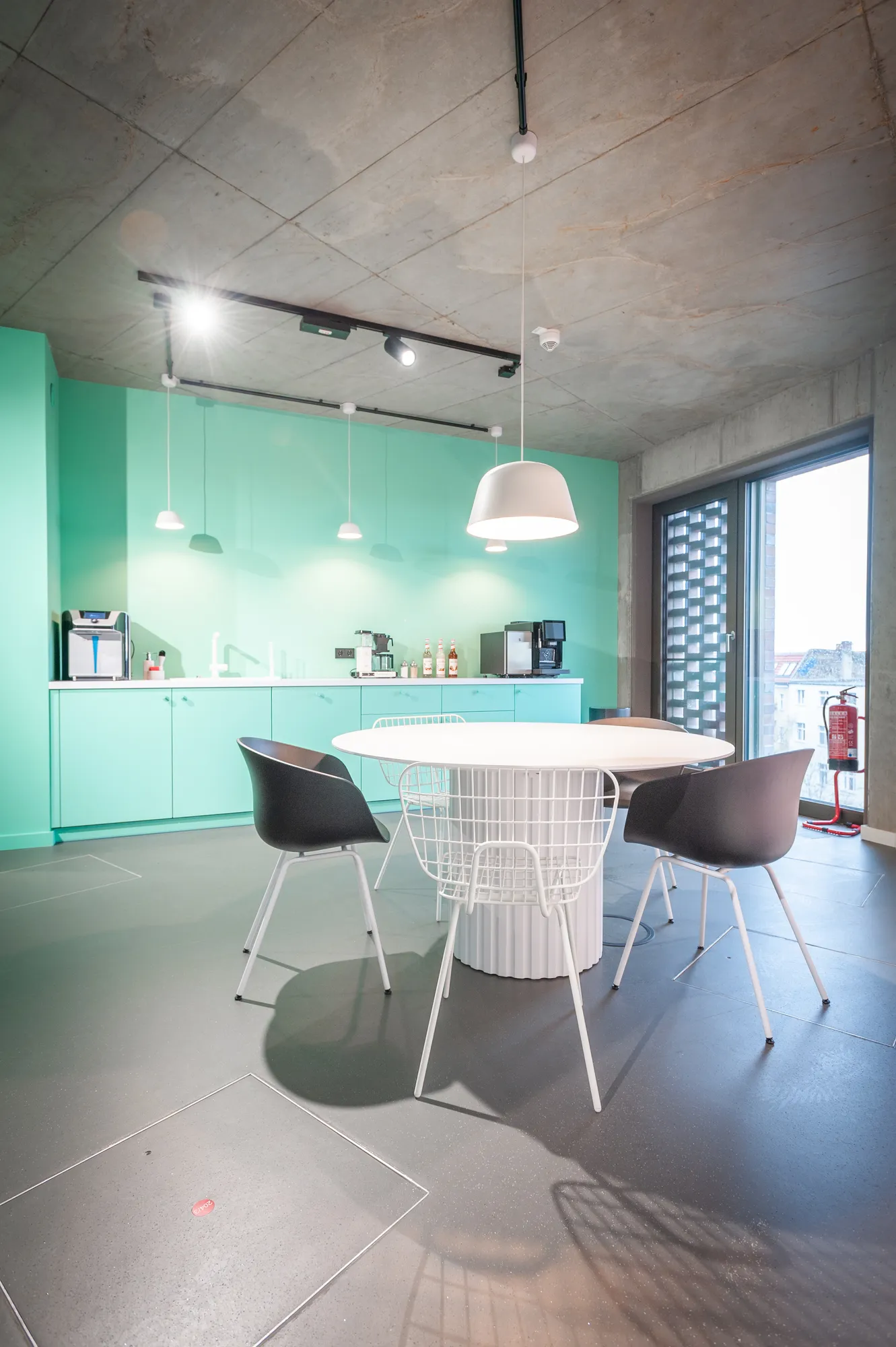 Mint-green kitchenette with round table, wire chair and city view through the window.