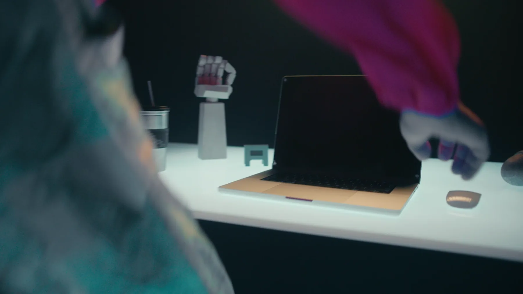 Over-the-shoulder view of a white desk with a laptop and a robotic hand sculpture, a second figure in a pink hoodie reaching into frame.