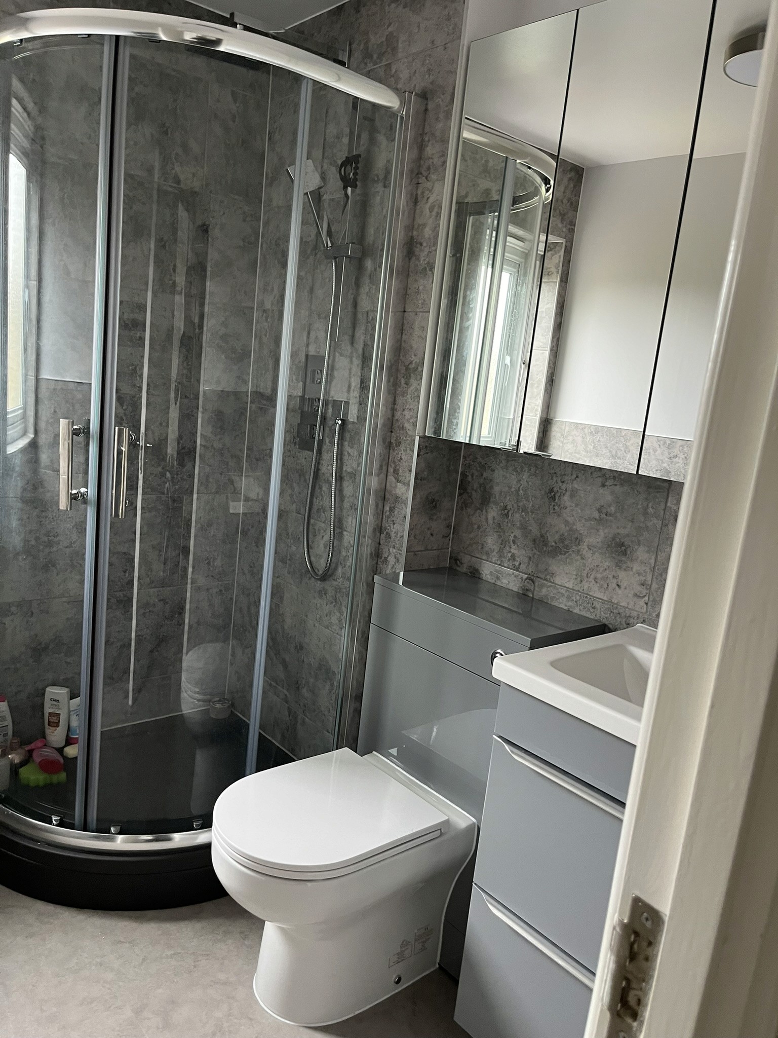 Bathroom with a curved glass shower enclosure, white toilet and grey vanity unit against stone-effect walls.