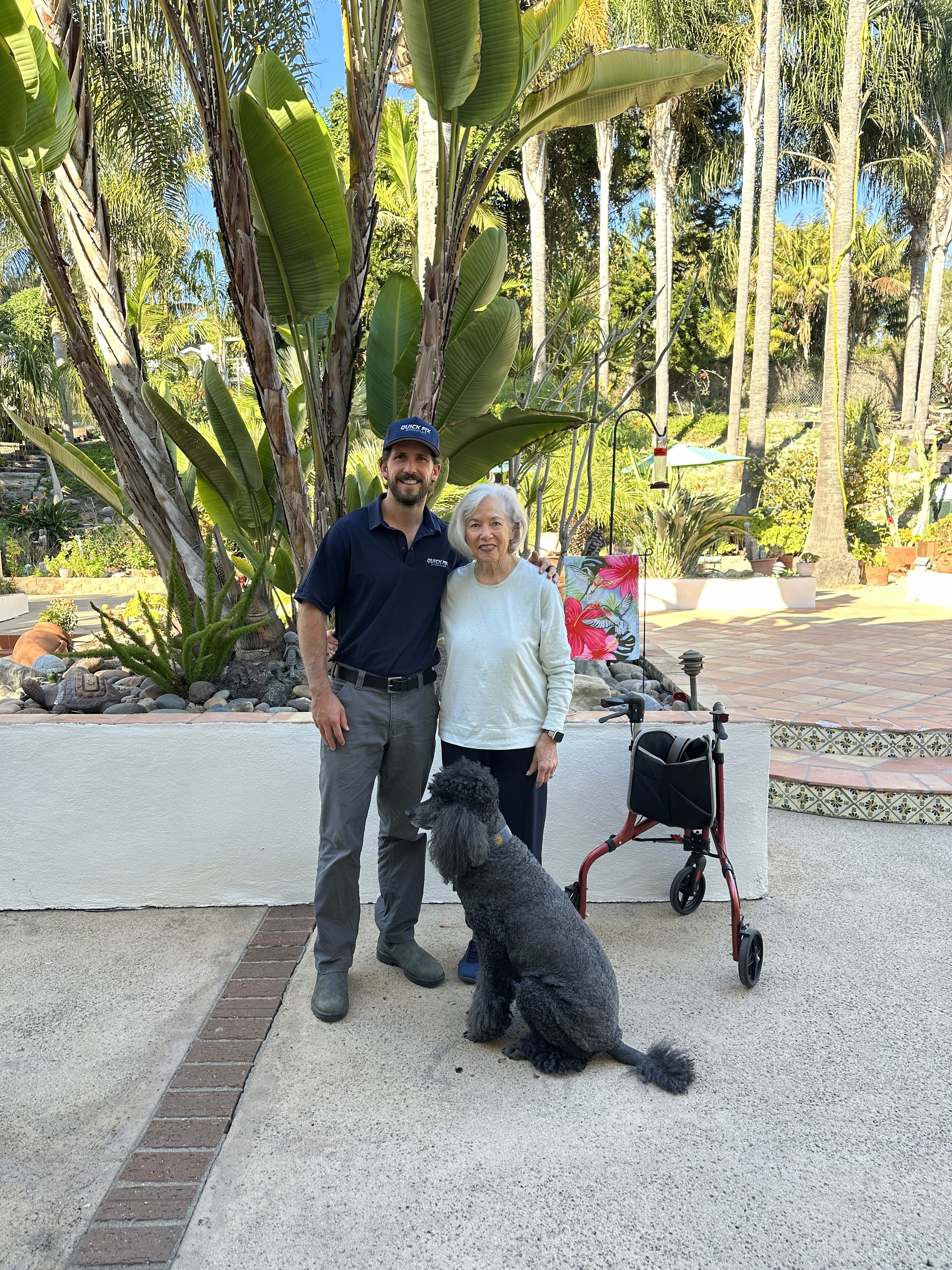 Christian, owner of Victory Sprinkler, sharing a friendly moment with customer Mary and her black poodle in her backyard after a successful sevice.