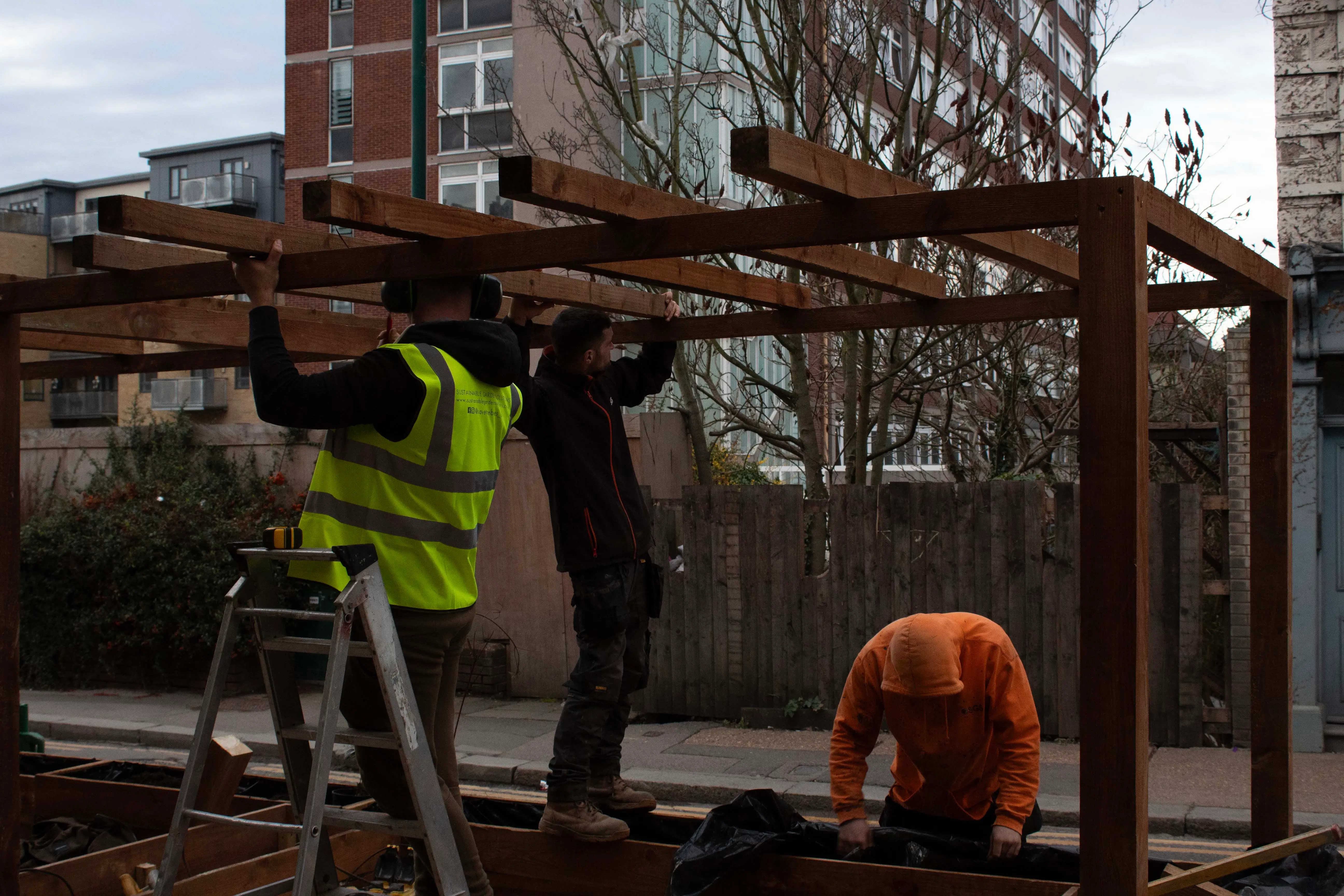 Two construction workers working on a wooden structure while using a ladder, with buildings in the background.