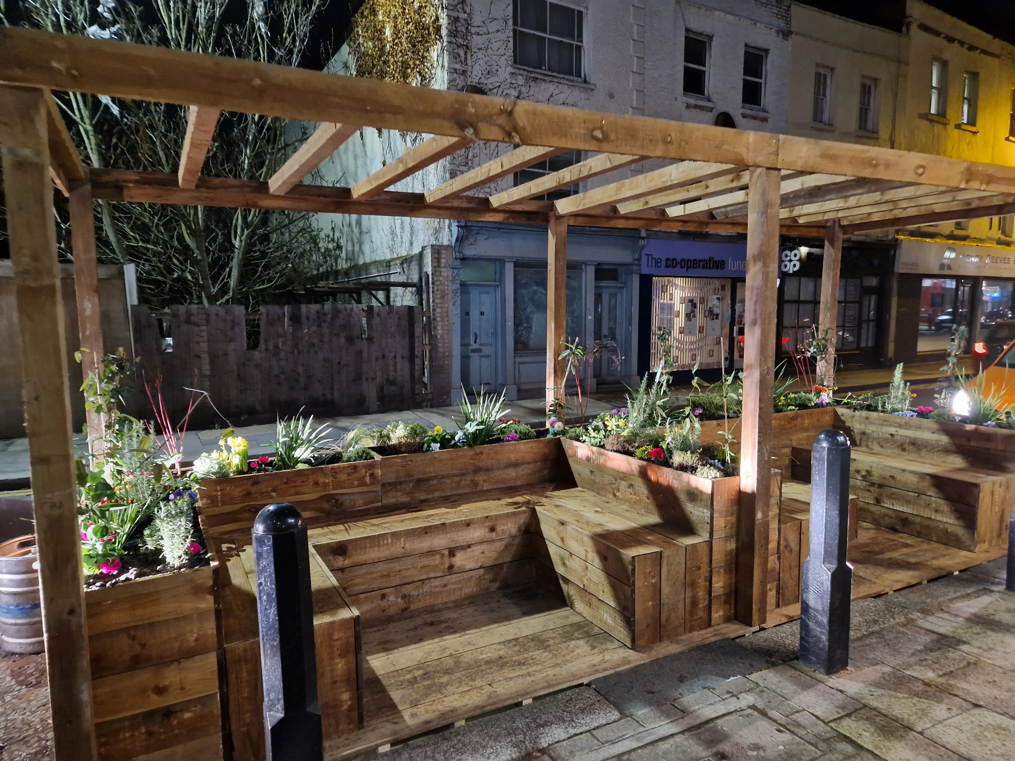 A well-lit outdoor seating area with wooden benches and greenery, set in an urban environment.