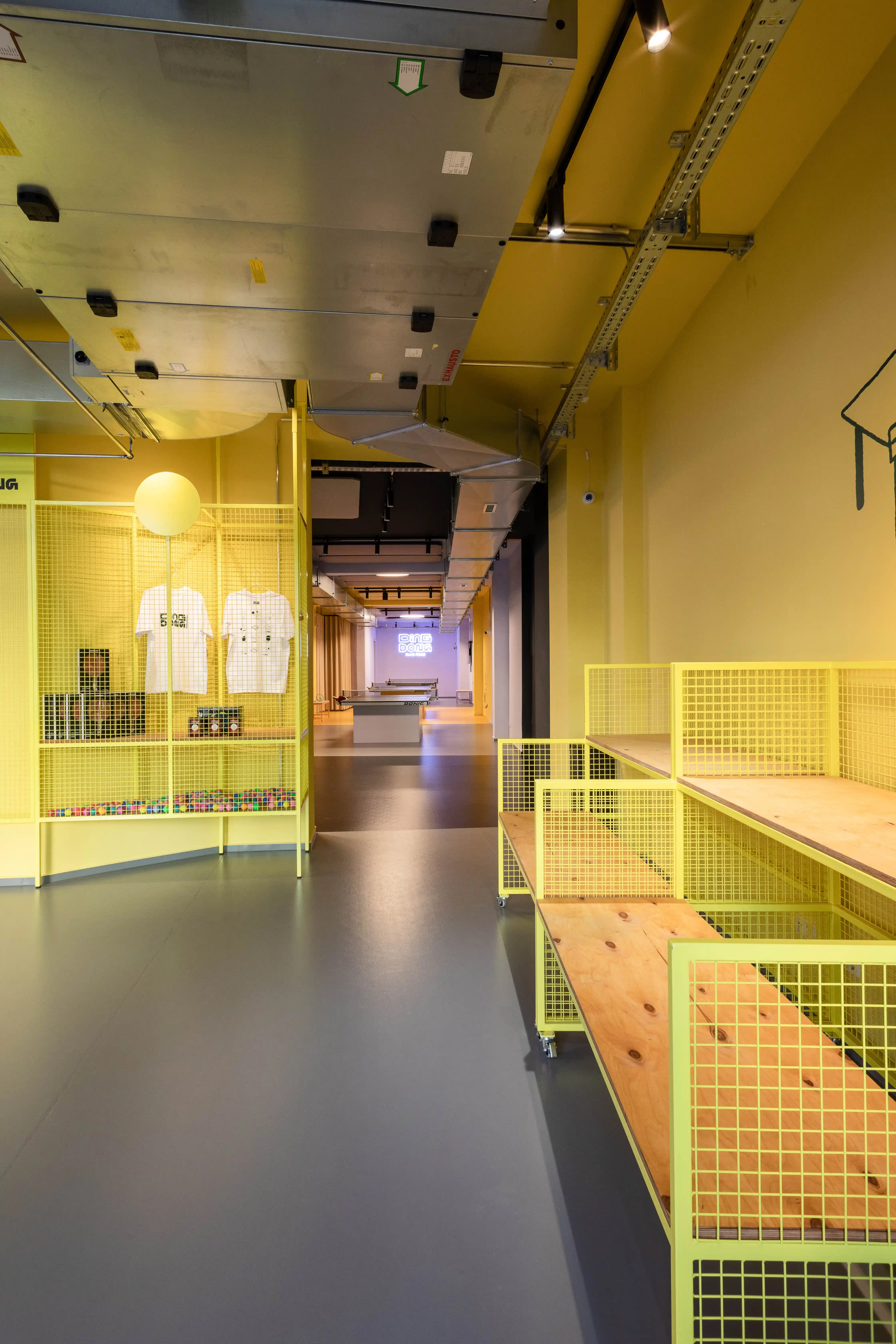 Corridor at DING DONG PING PONG lined with yellow wire-mesh shelving with wooden boards, leading toward the play hall with tables.