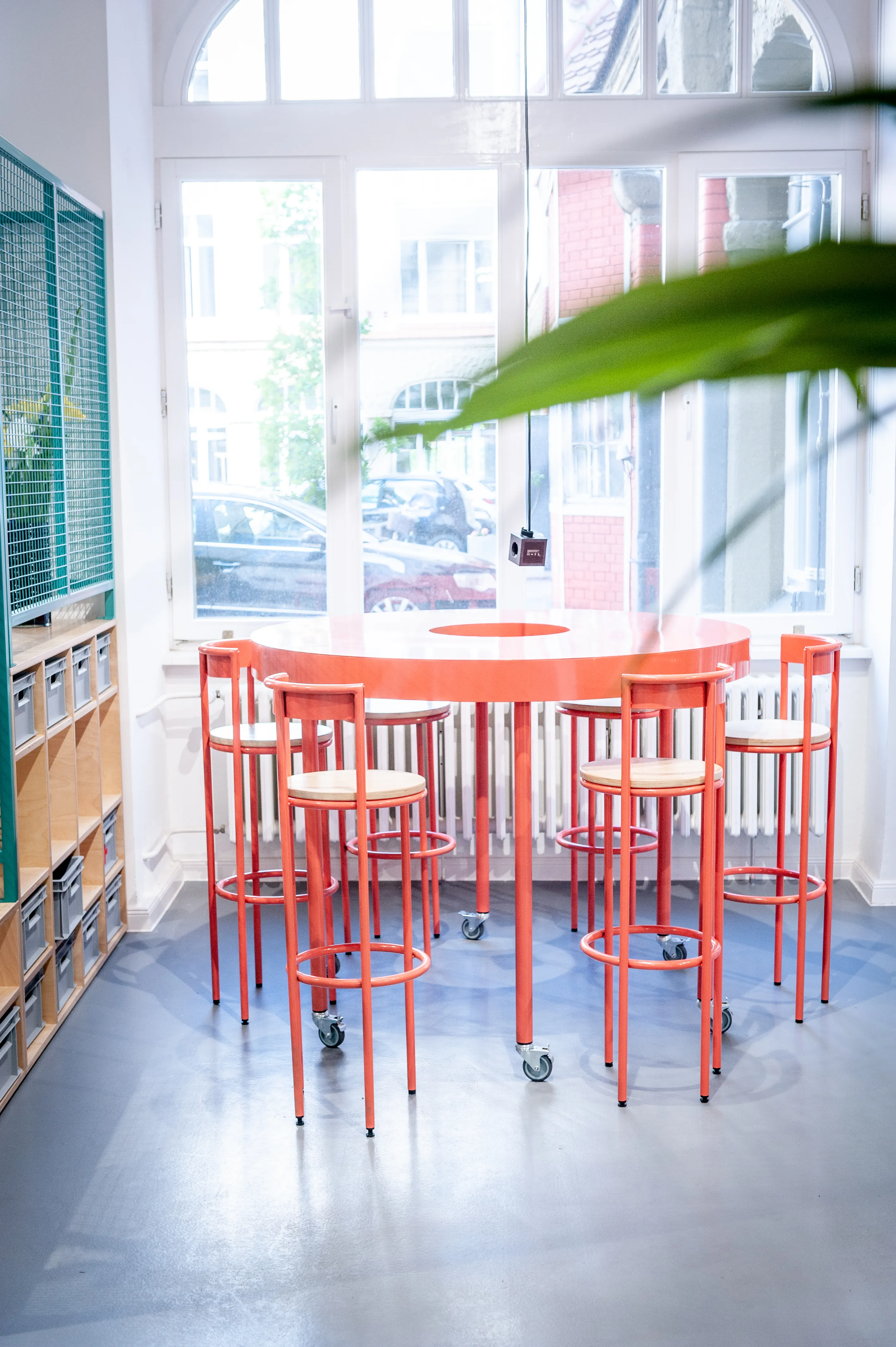Round red standing table with matching bar stools on castors at HEY CONTENT STUDIO, placed in front of large old-building windows.
