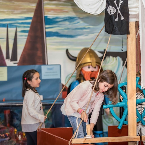 Two girls play on a pirate ship themed play area, with one girl pulling a rope and the other observing. A Viking statue is in the background.