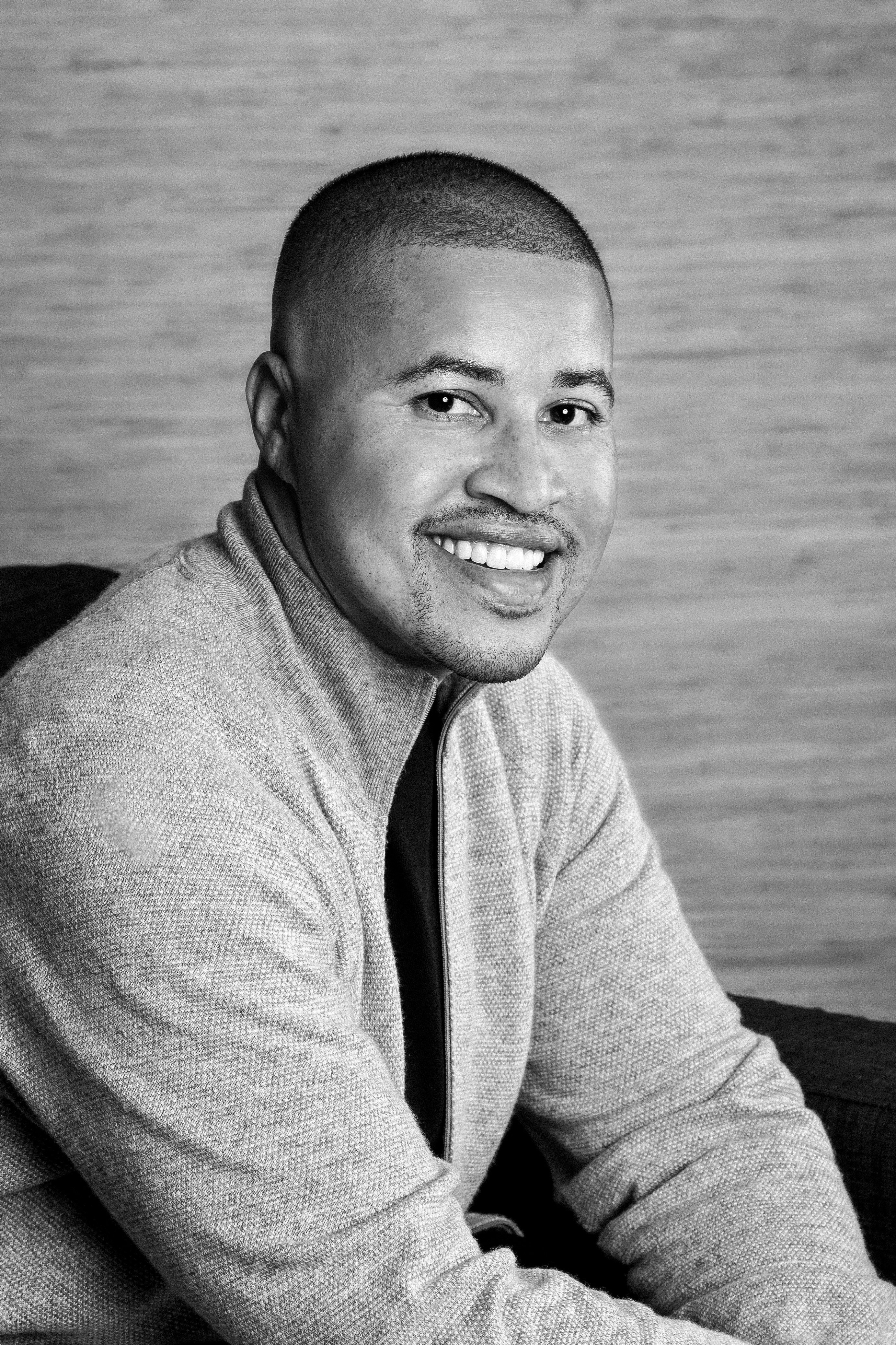 A young man with dark hair smiles at the camera, set against a modern indoor background.
