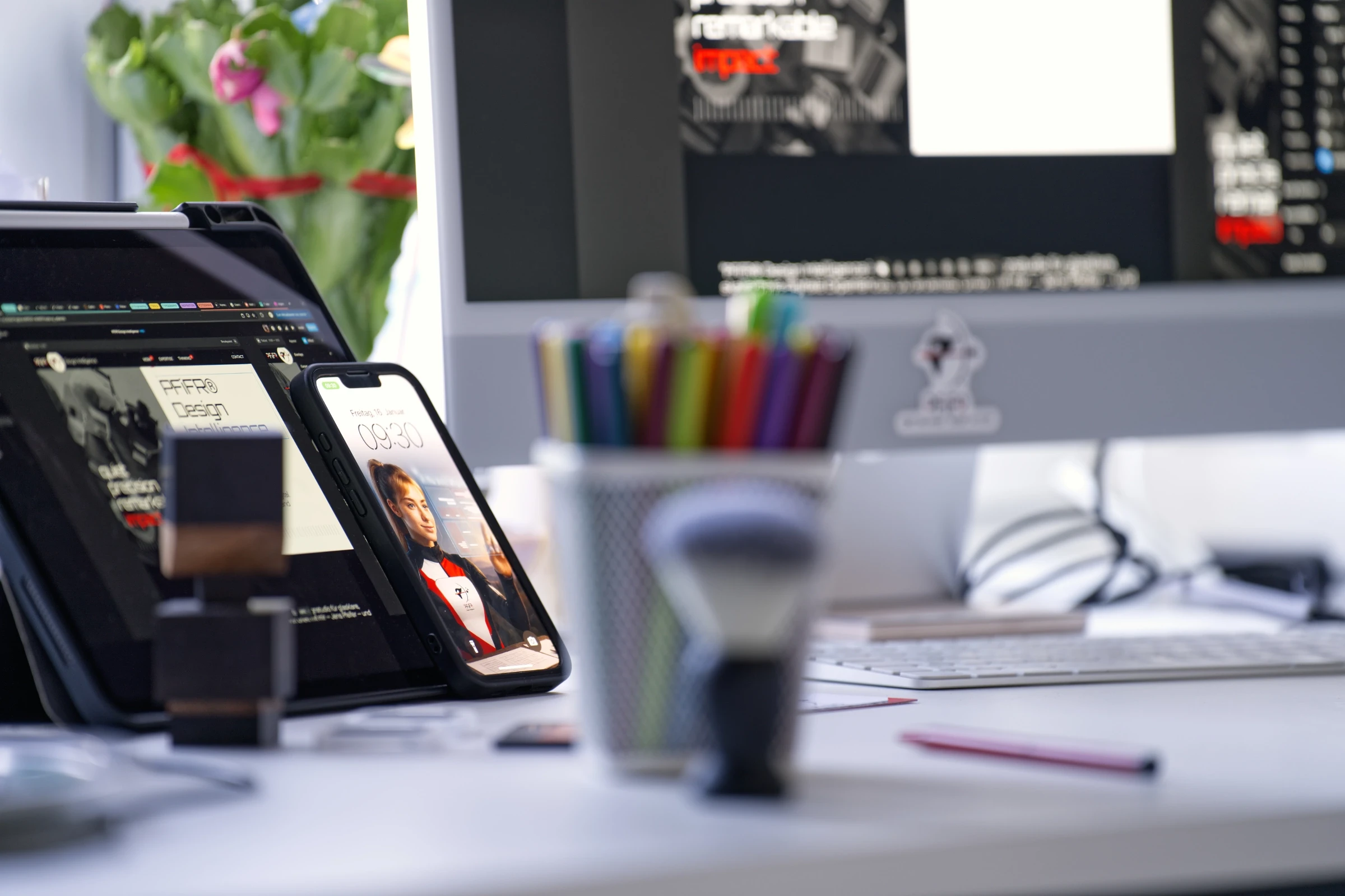 Detailaufnahme eines iPads und iPhones auf einem Design-Schreibtisch, beide zeigen die Website „PFiFR Design Intelligence“, im Vordergrund liegen Zeichenstifte und Werkzeuge, im Hintergrund ein Desktop-Monitor.