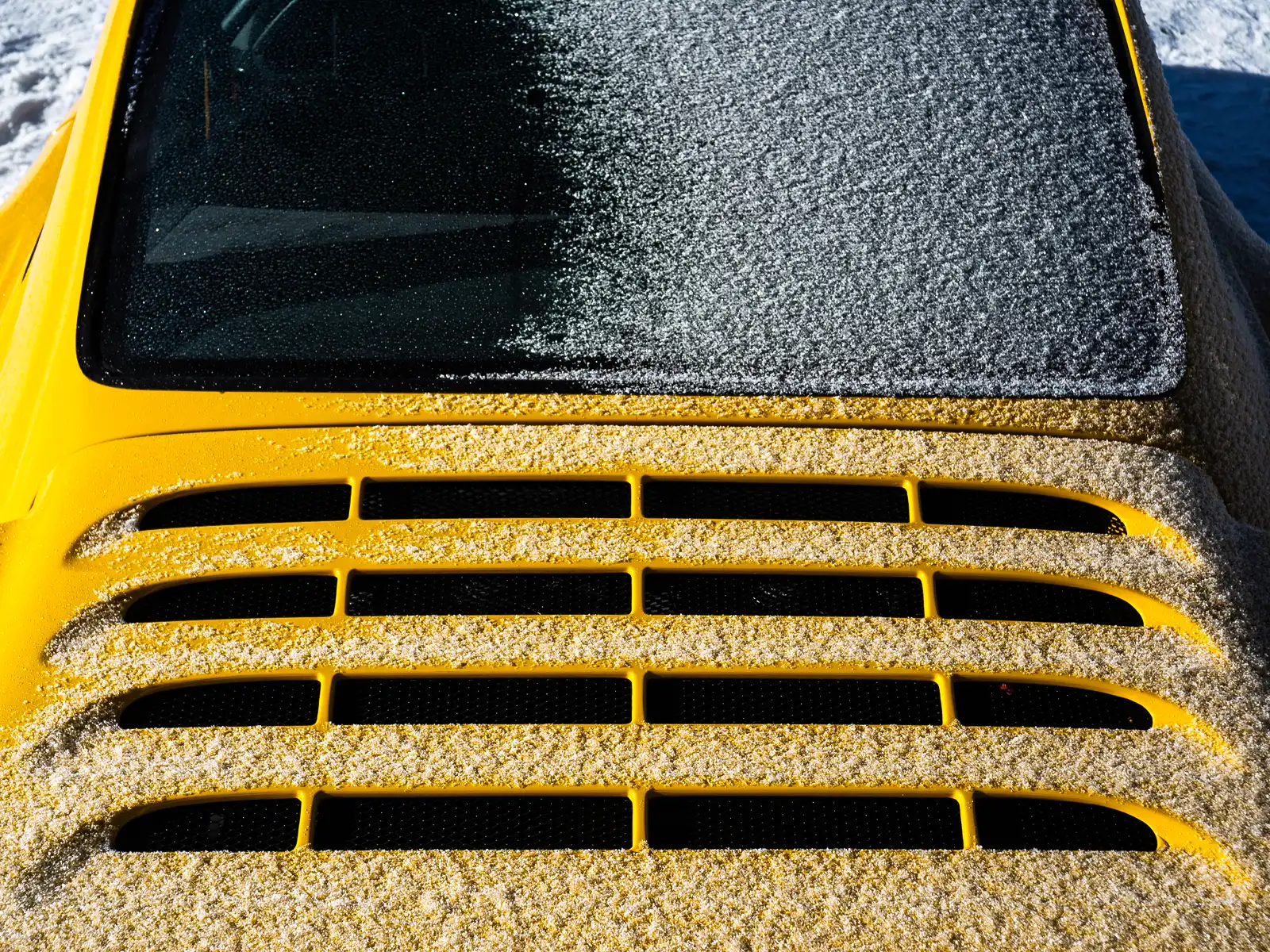 The rear window and engine cover of a yellow Porsche parked outside, concealed beneath a blanket of white snow.