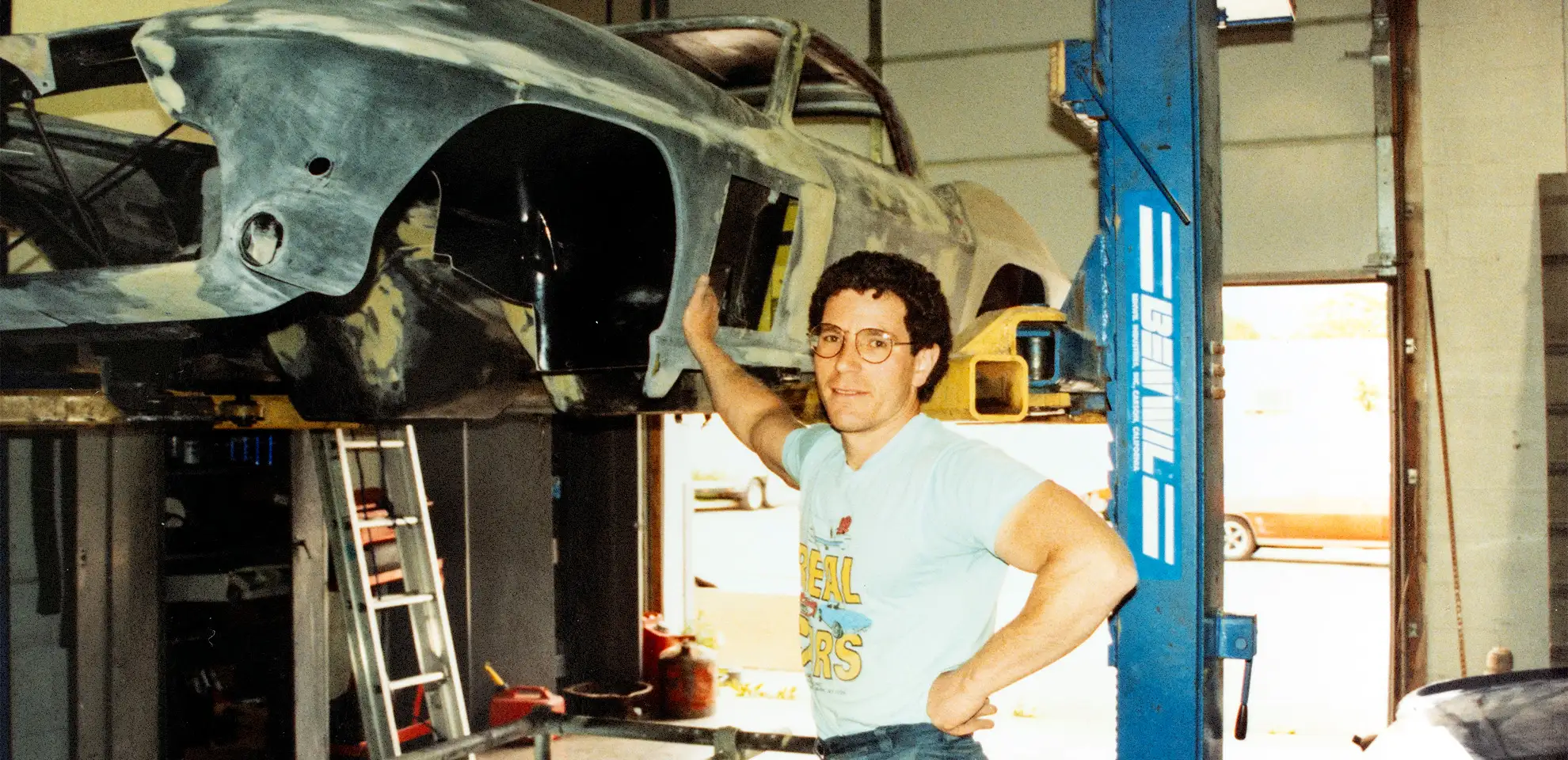 A man in glasses stands beside a classic car frame on a lift in a workshop.