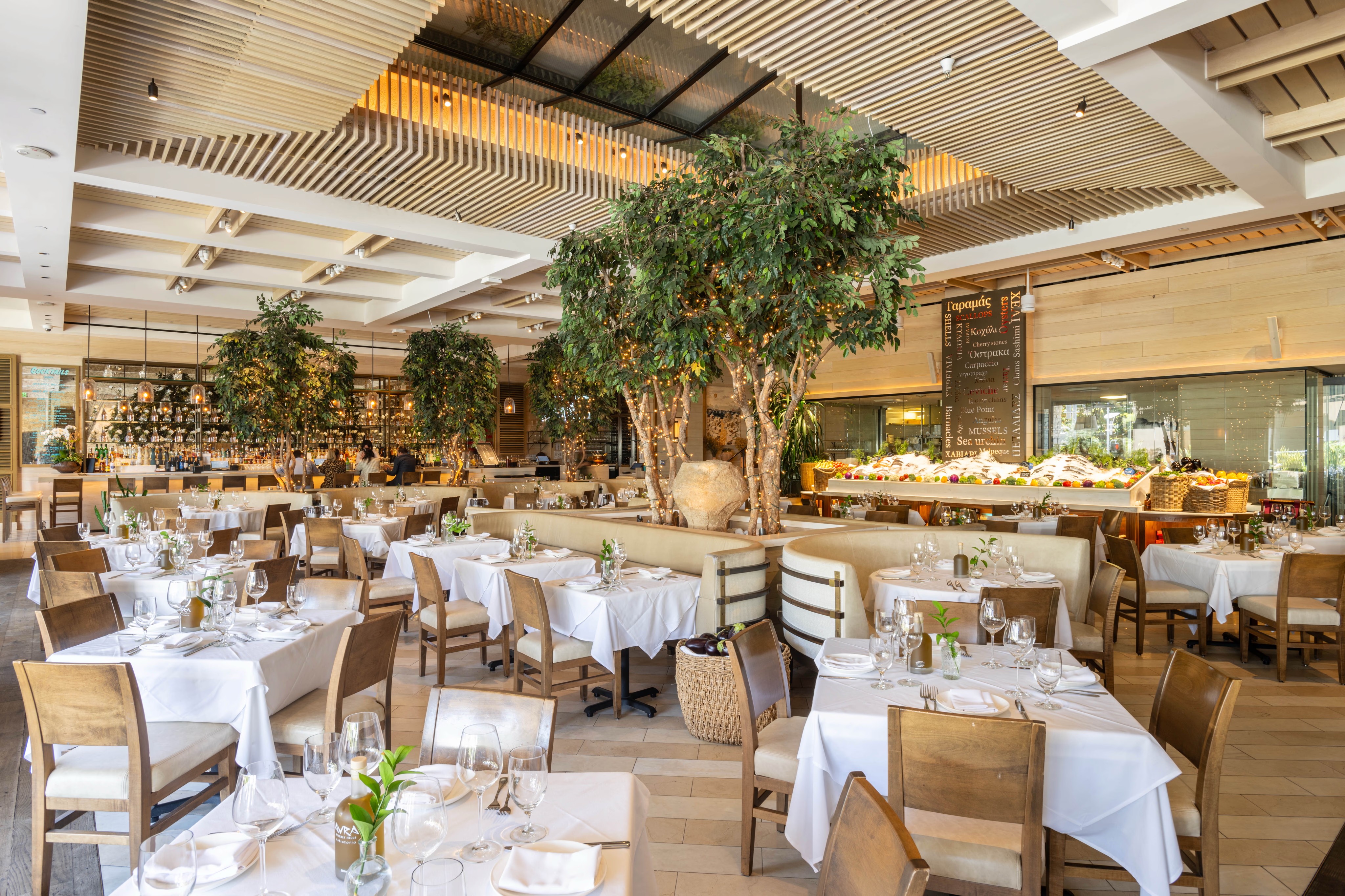 A spacious restaurant interior with white tablecloths, wooden accents, and decorative trees.