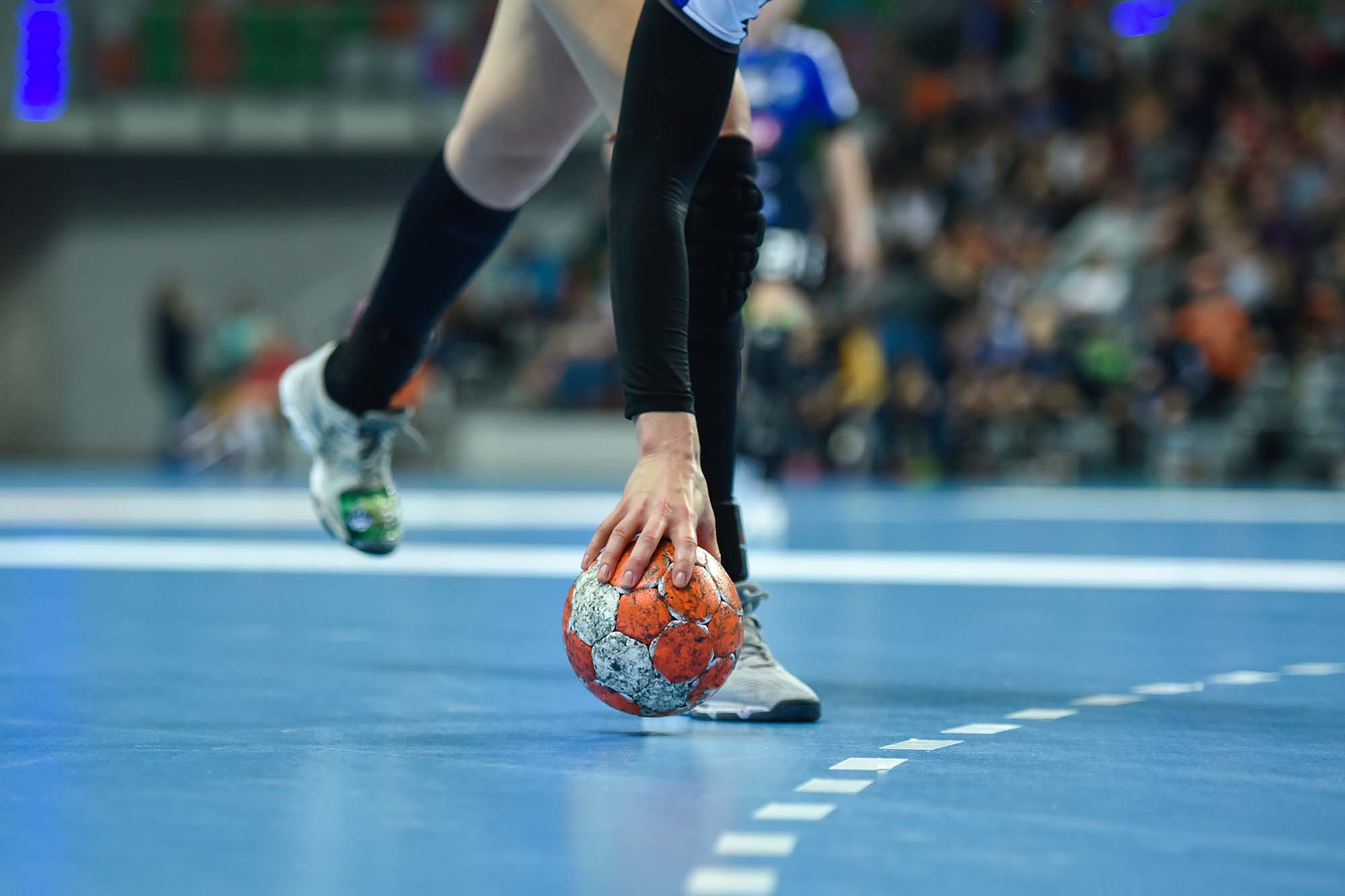 Close-up of a handball player picking up a worn red ball off the blue court floor during a match.