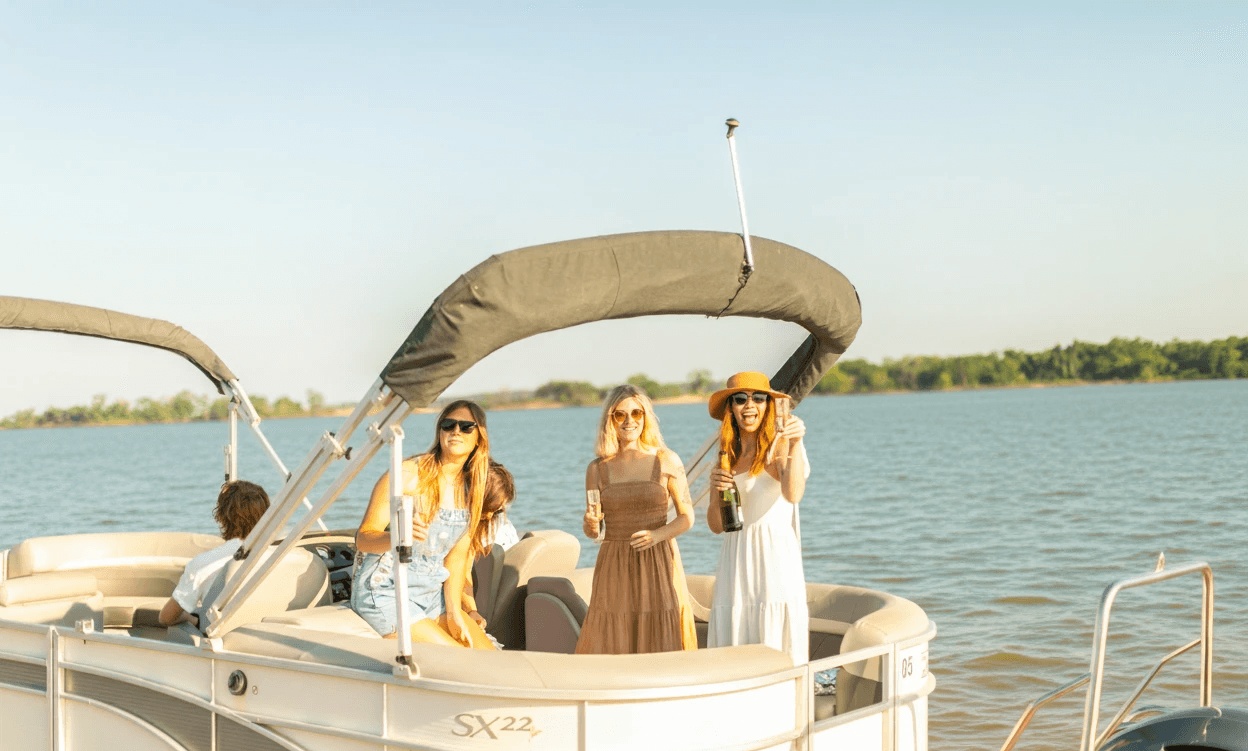 A group of people enjoy a sunny day on a pontoon boat with drinks in hand, set against the backdrop of a serene lake and clear blue sky.