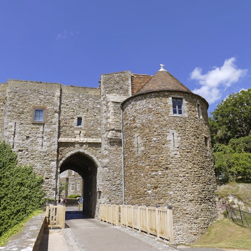 ทางเข้าสู่ปราสาทหินที่มีหอคอยกลม สะพานไม้ และต้นไม้ล้อมรอบ ใต้ท้องฟ้าสีฟ้าใส