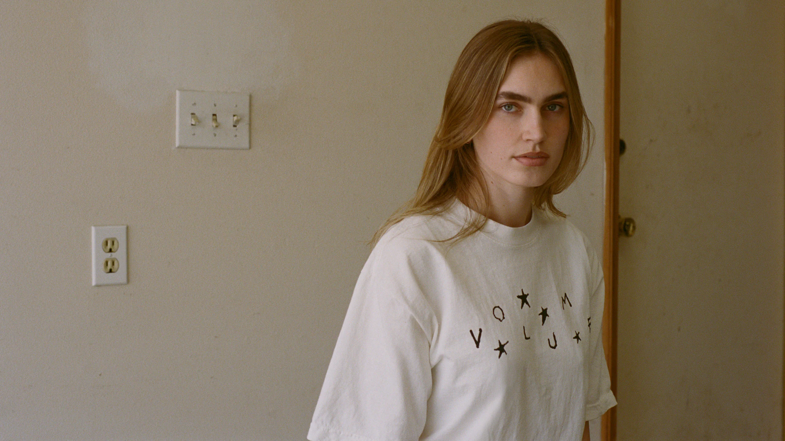 A model wears the Stars + Scribbles T-shirt by Volume LWH, standing indoors against a plain wall with visible outlets for the Vol. 1 “Garage” campaign.