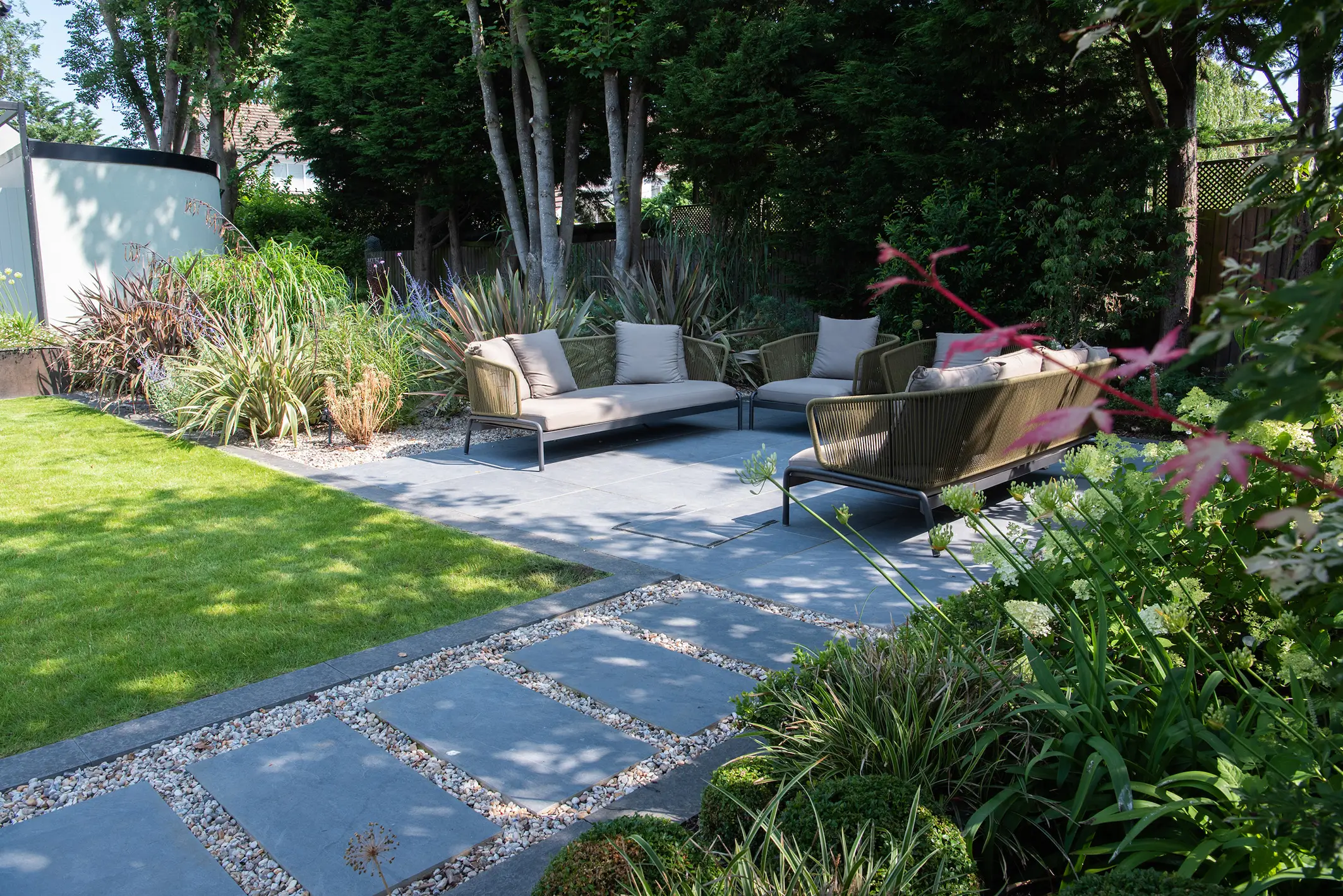 A landscaped garden featuring a stone pathway, seating area, and greenery in a sunny setting.