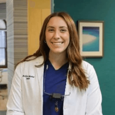 Alt tag: Portrait of Dr. Erin Madar smiling in a dental office, wearing blue scrubs and a white lab coat with dental loupes around her neck.