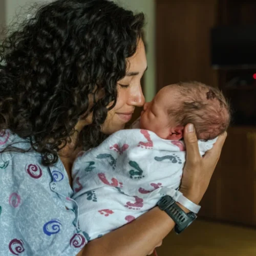 Mother gently cradling her swaddled newborn baby at home, preparing for a medical procedure in Melbourne's northern suburbs.