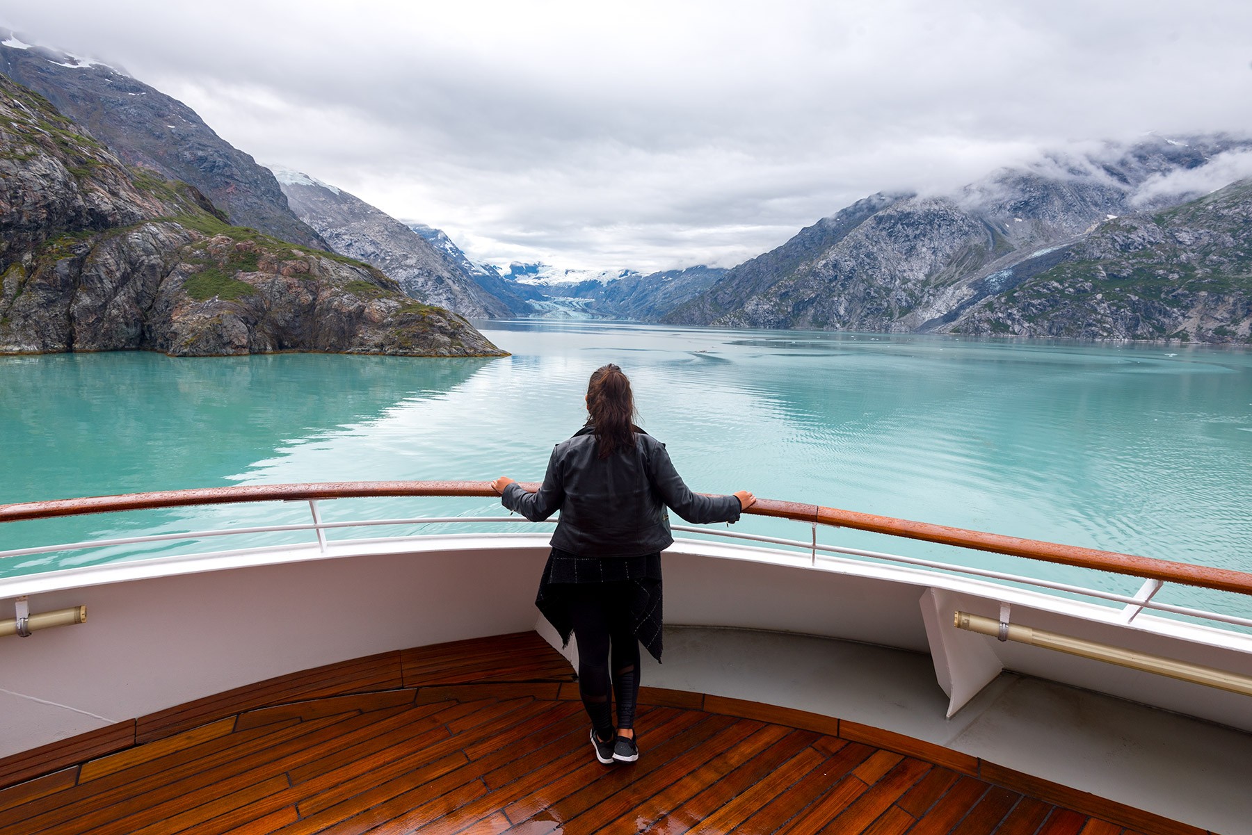 A person stands on a wooden cruise ship deck with a railing, looking out over a turquoise glacial lake and mountains.